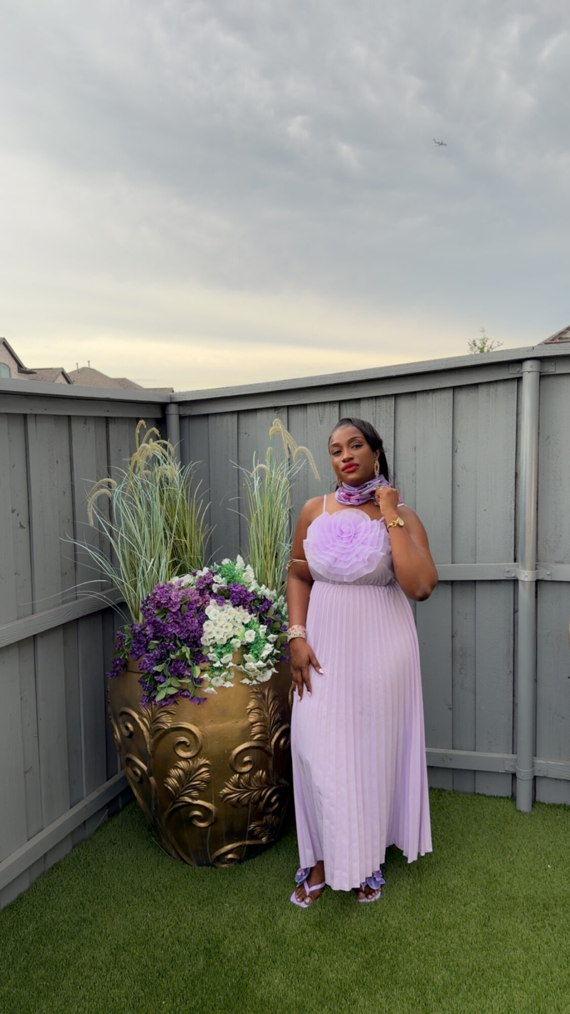 Woman in a long, light purple dress standing next to a large gold decorative planter with purple and white flowers, outdoors on grass.
