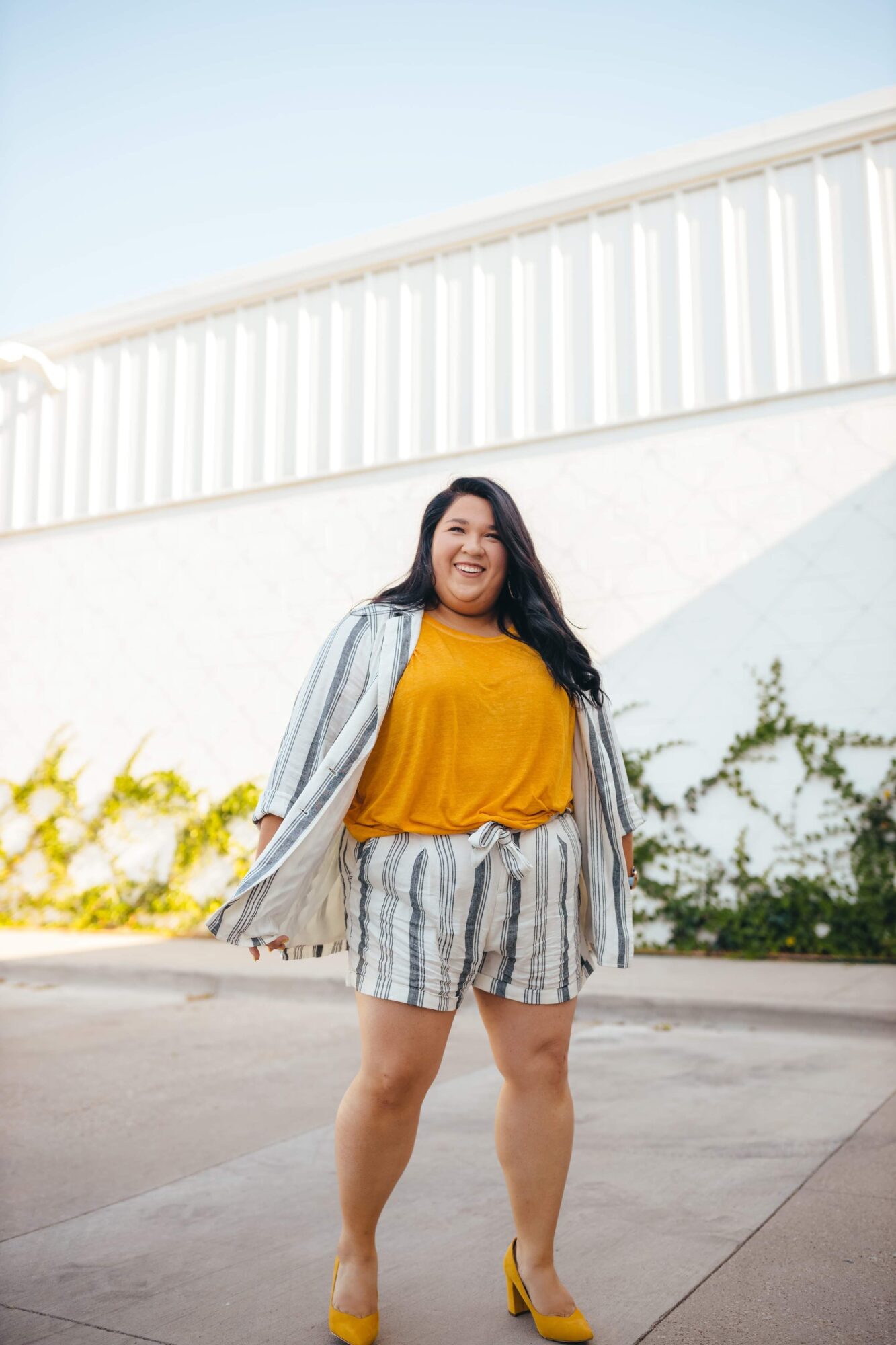 Woman standing outdoors, smiling, wearing a yellow top, striped shorts, and yellow heels, with a white building and greenery behind her.