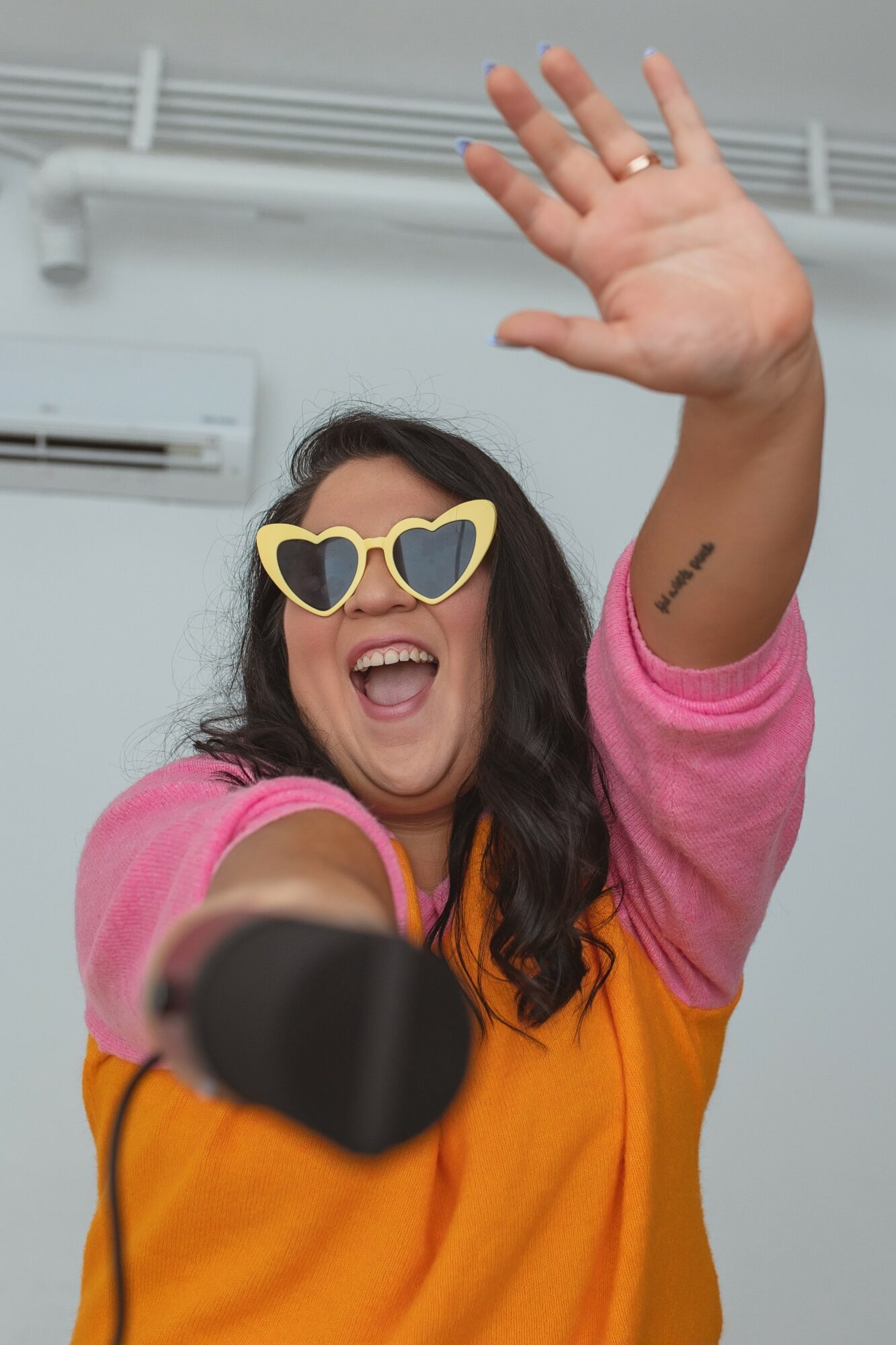 Woman wearing heart-shaped sunglasses and a pink and orange shirt, smiling and waving with her hand raised.