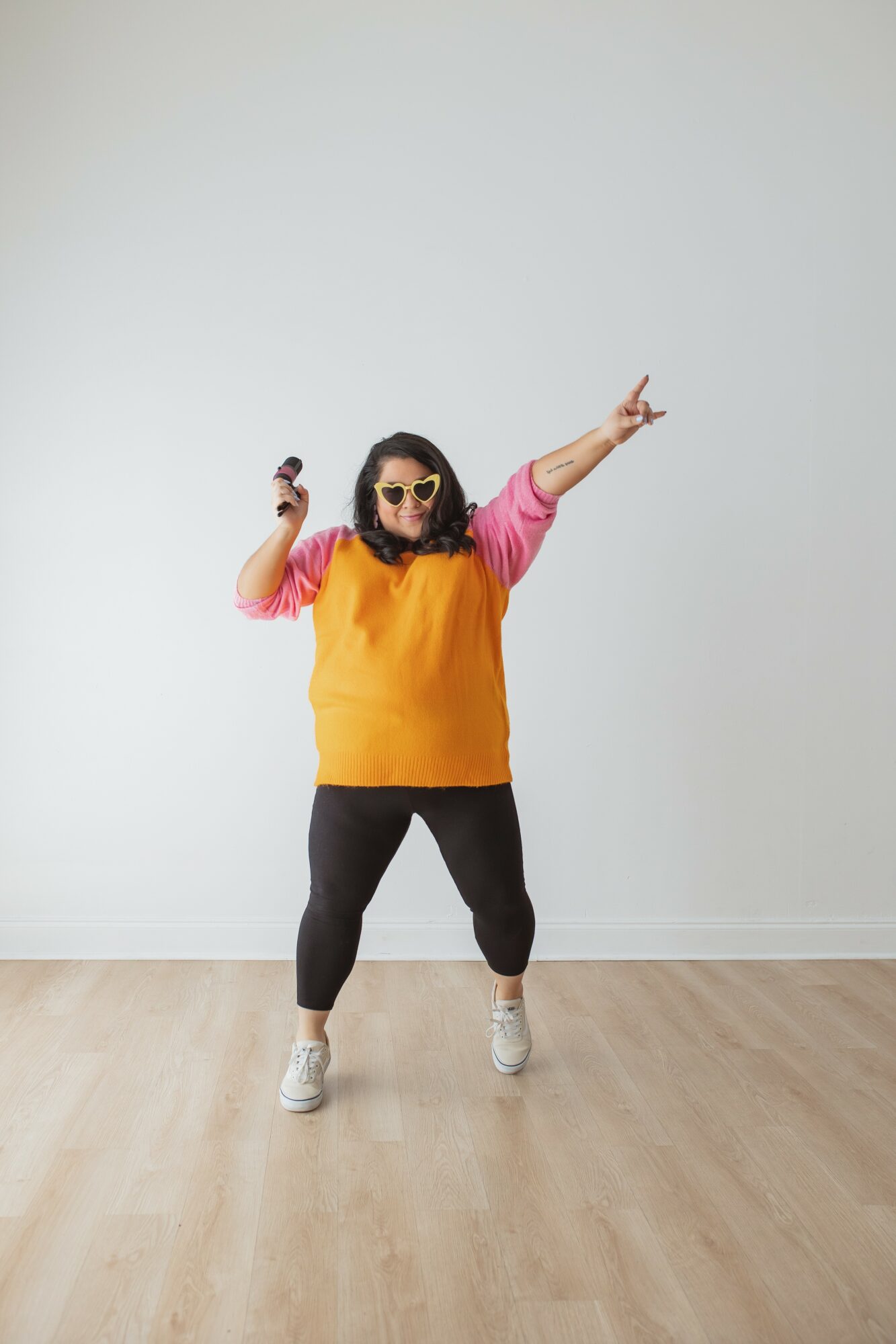 Person dancing with sunglasses, wearing a yellow and pink top, black pants, and white shoes, against a plain white wall.