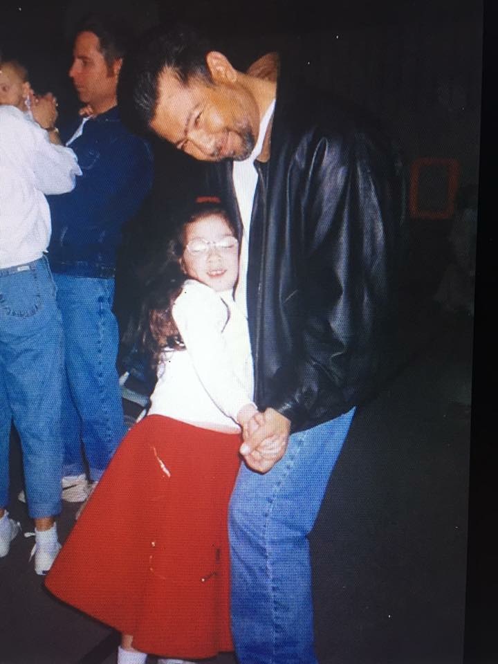 A man and a young girl hold hands and smile, with other people in the background. The man wears a leather jacket.