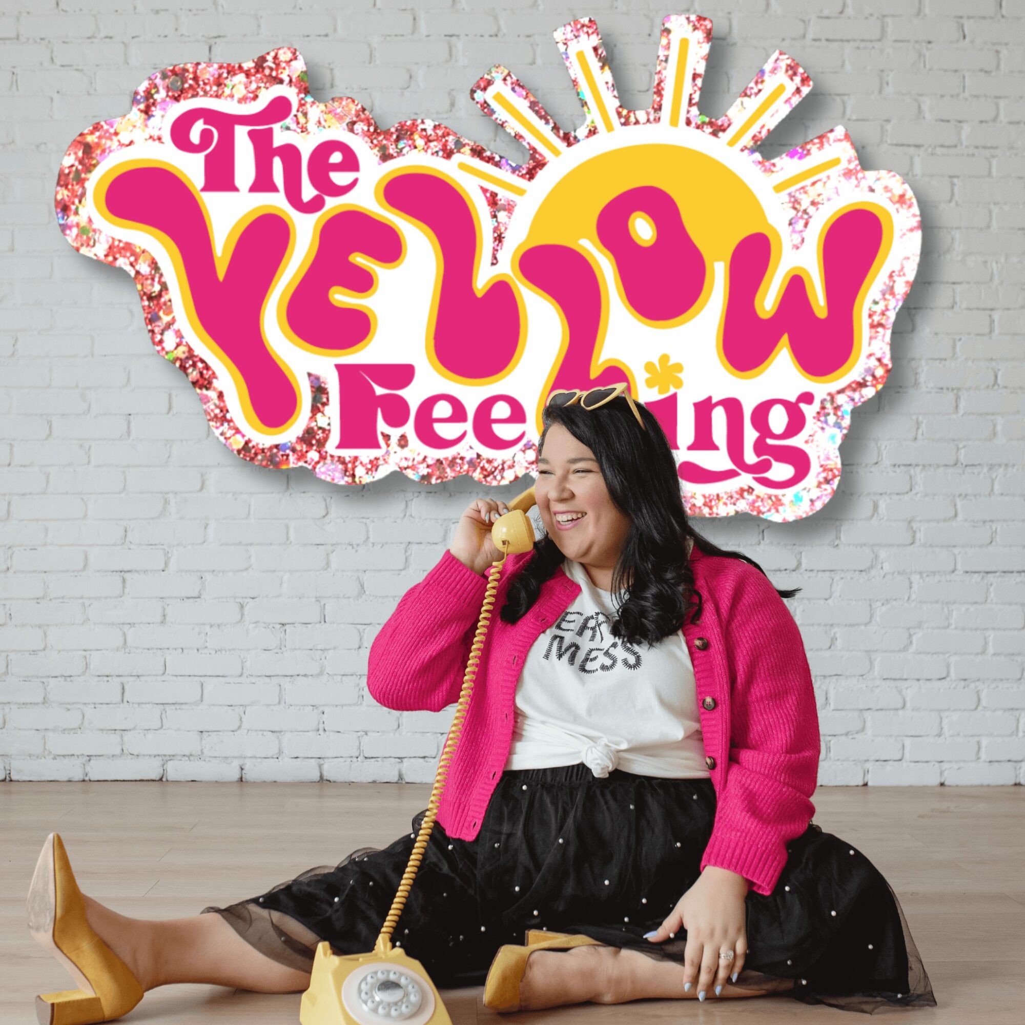Woman sitting on floor with a yellow phone, smiling, in front of a colorful sign that says 'The Yellow Feeling'.