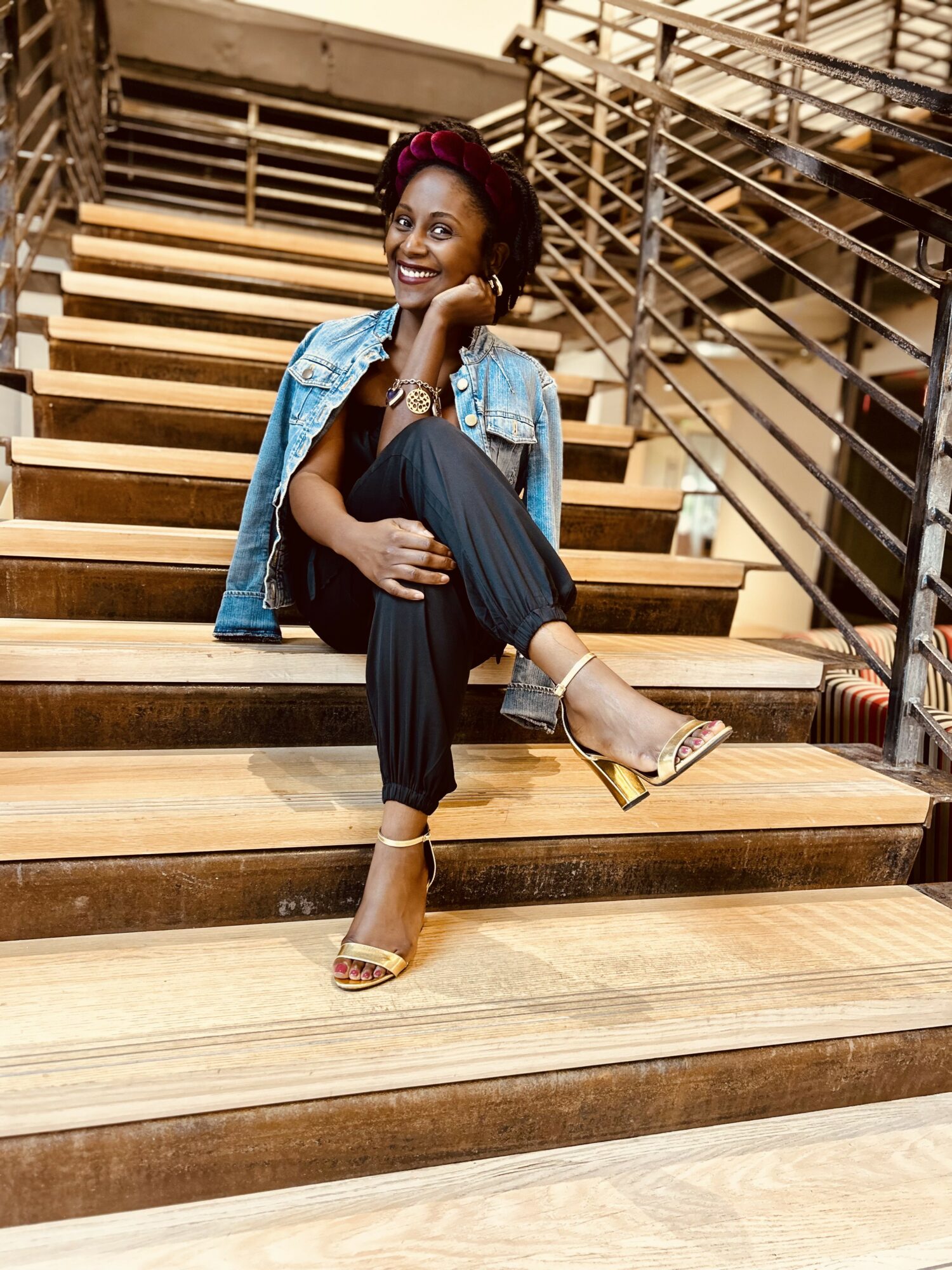Woman sitting on wooden stairs, smiling, wearing a denim jacket, black pants, and high-heeled sandals, with metal railings in background.