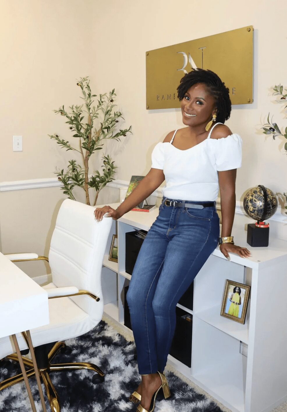 Woman standing in a room with plants, white furniture, and a gold wall sign, smiling at the camera.