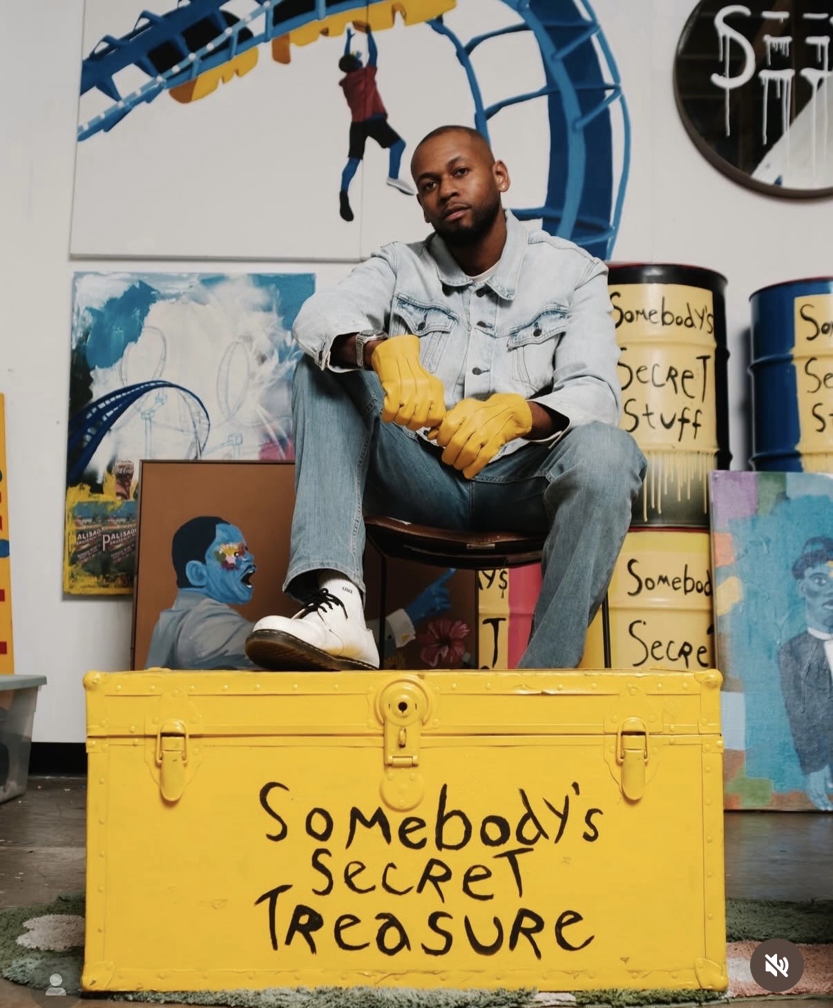Man sitting on a yellow box labeled 'Somebody's Secret Treasure' surrounded by artwork and storage containers.