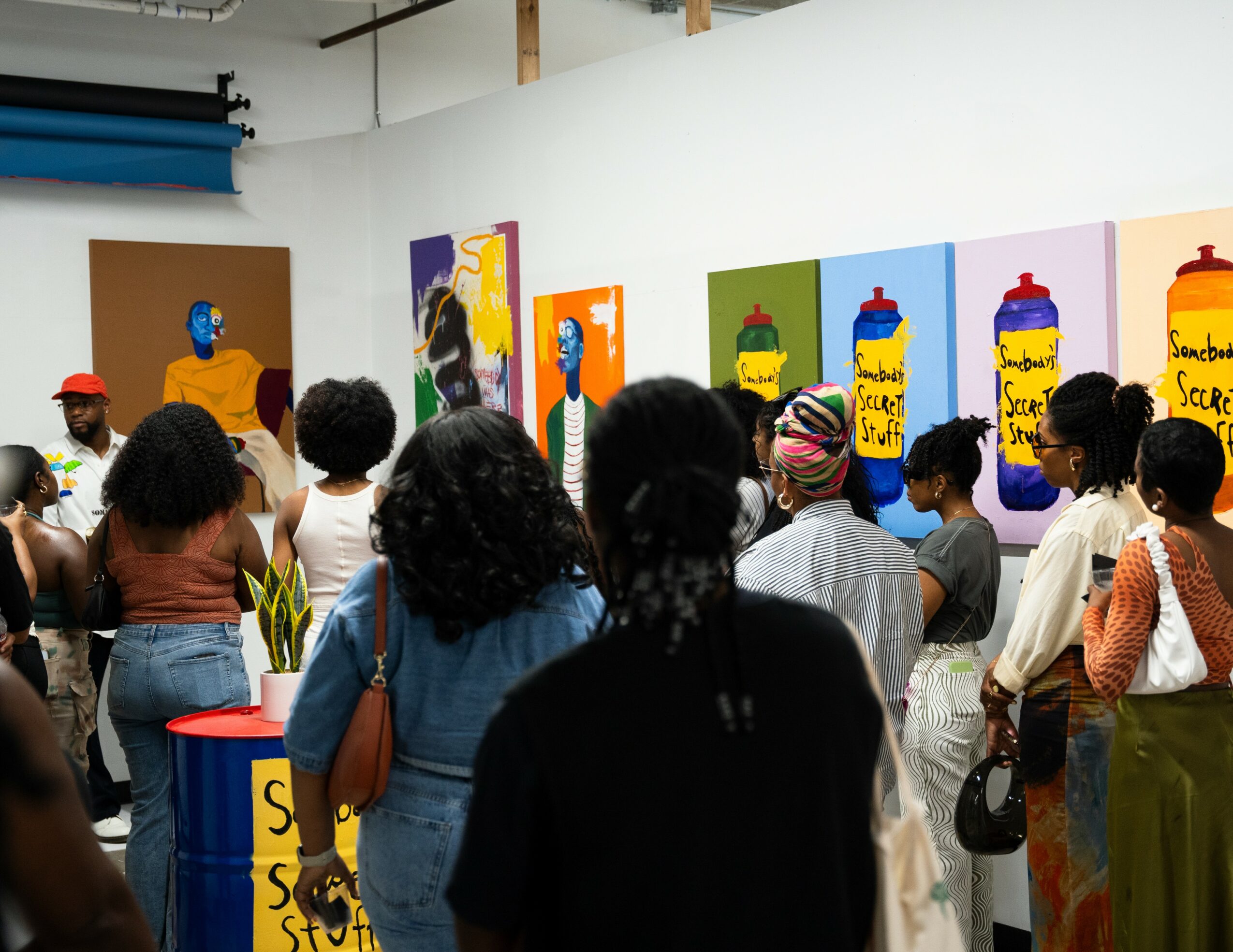 Group of people viewing colorful artwork on gallery wall, some wearing hats and carrying bags, in an indoor art space.