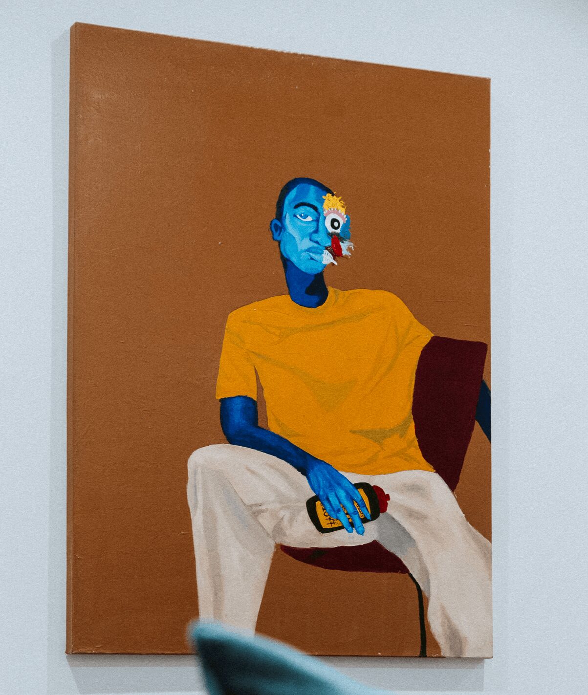 Painting of a blue-skinned person with a yellow shirt sitting on a chair, holding a remote, against a brown background.