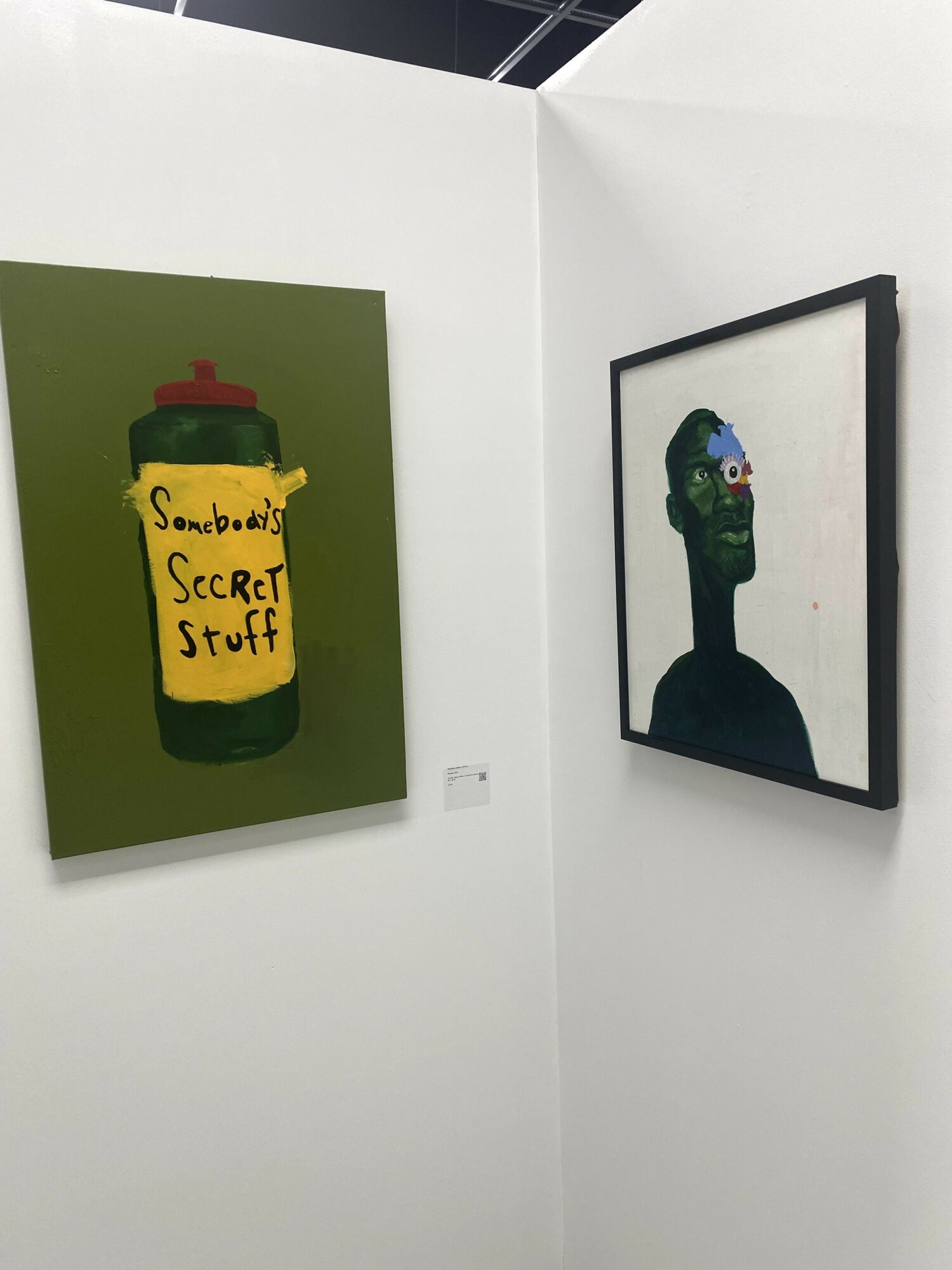 Two framed artworks on white walls; one depicts a yellow container with a red lid and a note reading 'Secret Stuff,' the other shows a stylized green face with large eyes and a dark background.