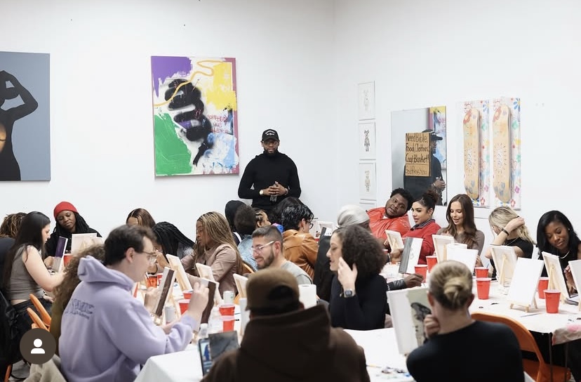 Group of people seated at tables in an art gallery, listening to a speaker, with colorful artwork on the walls.
