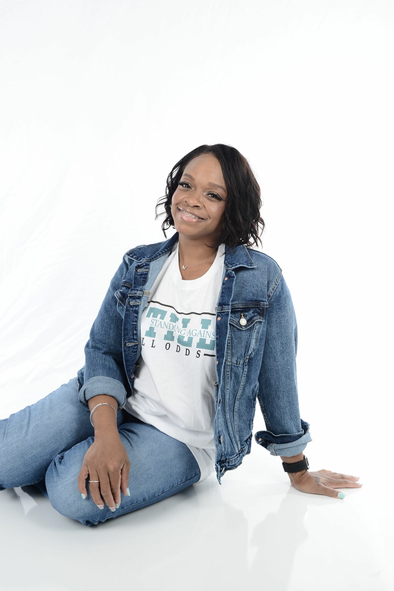 Woman sitting on the floor with legs crossed, smiling, wearing a denim jacket and jeans, against a plain white background.