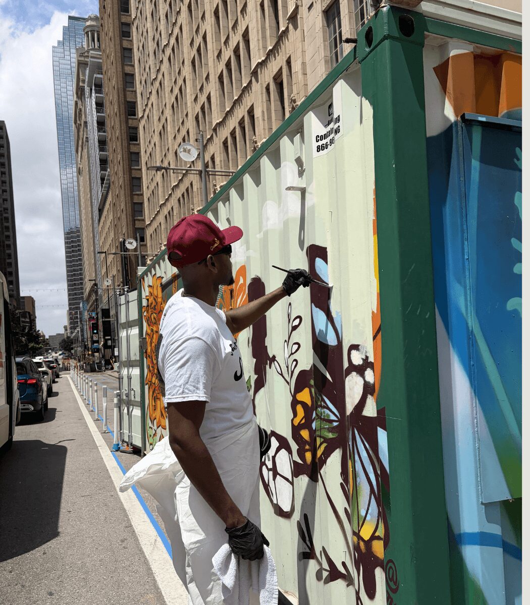 Person painting graffiti on a large container in an urban street, tall buildings in the background.