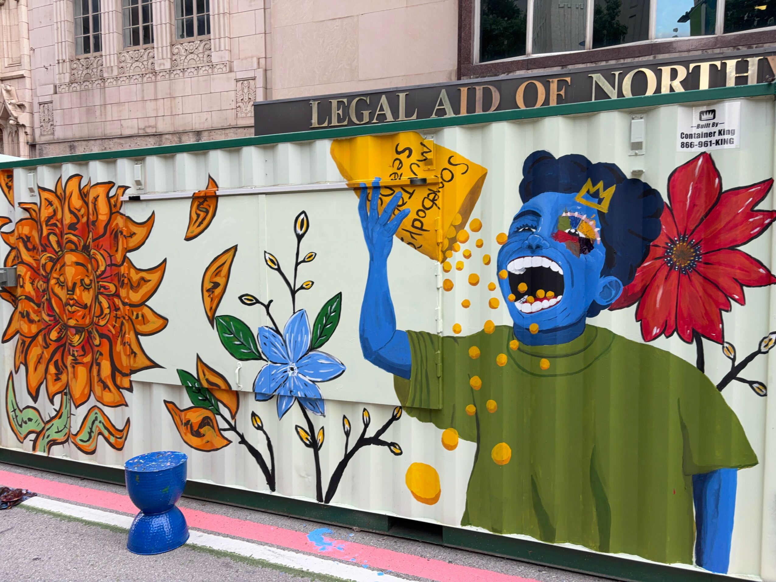 Colorful mural featuring a blue creature with a crown, surrounded by large flowers and leaves, with a sign reading 'LEGAL AID OF NORTH'.