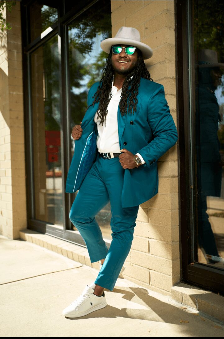 Person in teal suit, white shirt, white sneakers, wide-brimmed hat, sunglasses, standing outside near glass door, smiling.