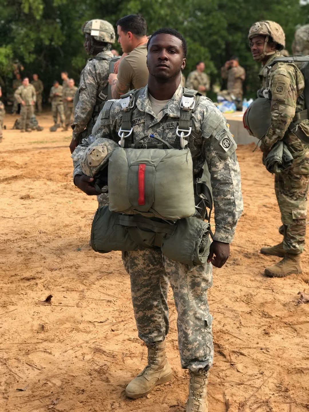 Soldier in camouflage uniform carrying gear, standing outdoors with other soldiers in background.
