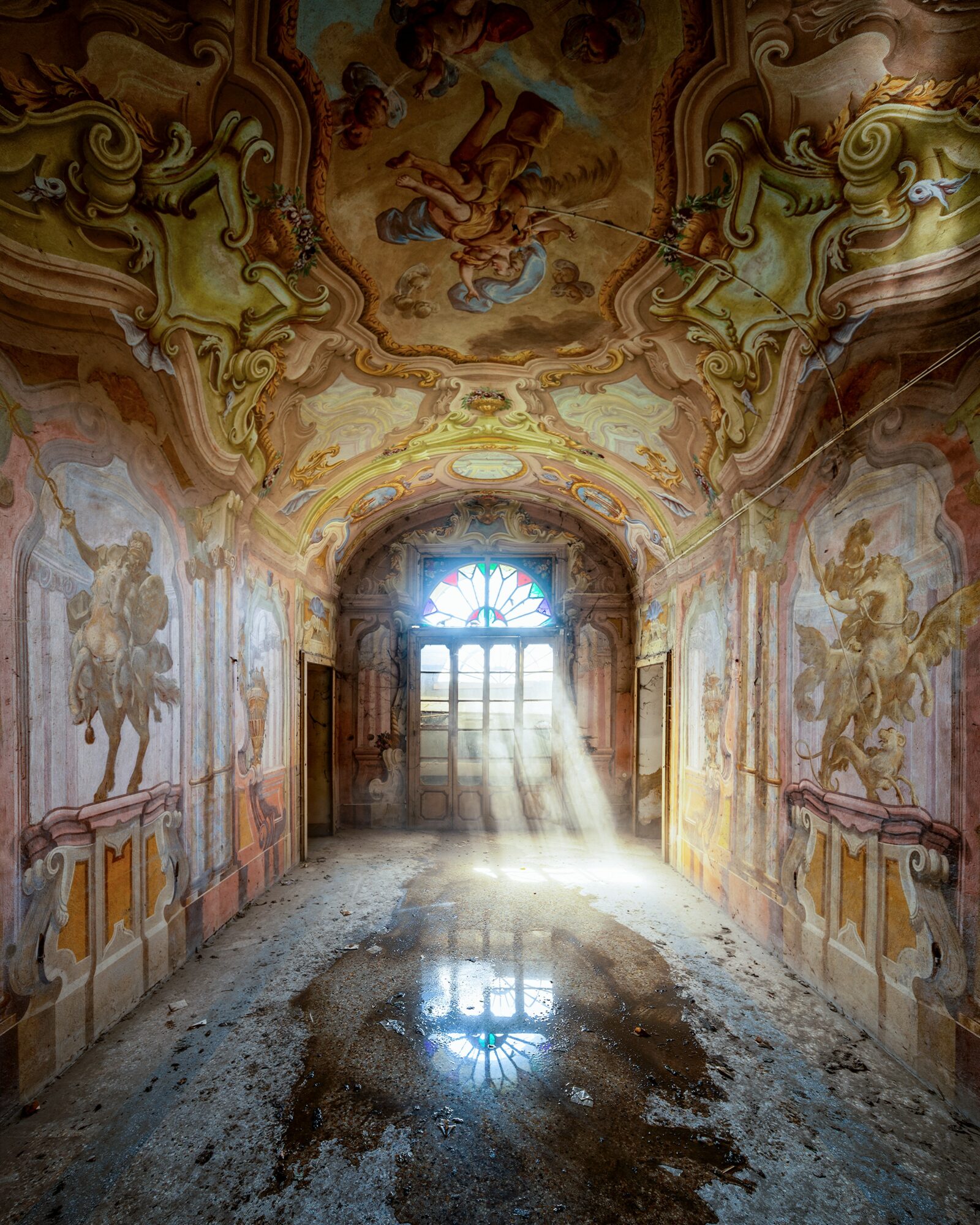 Long corridor with ornate ceiling paintings and sunlight streaming through a window at the end, reflecting on the floor.