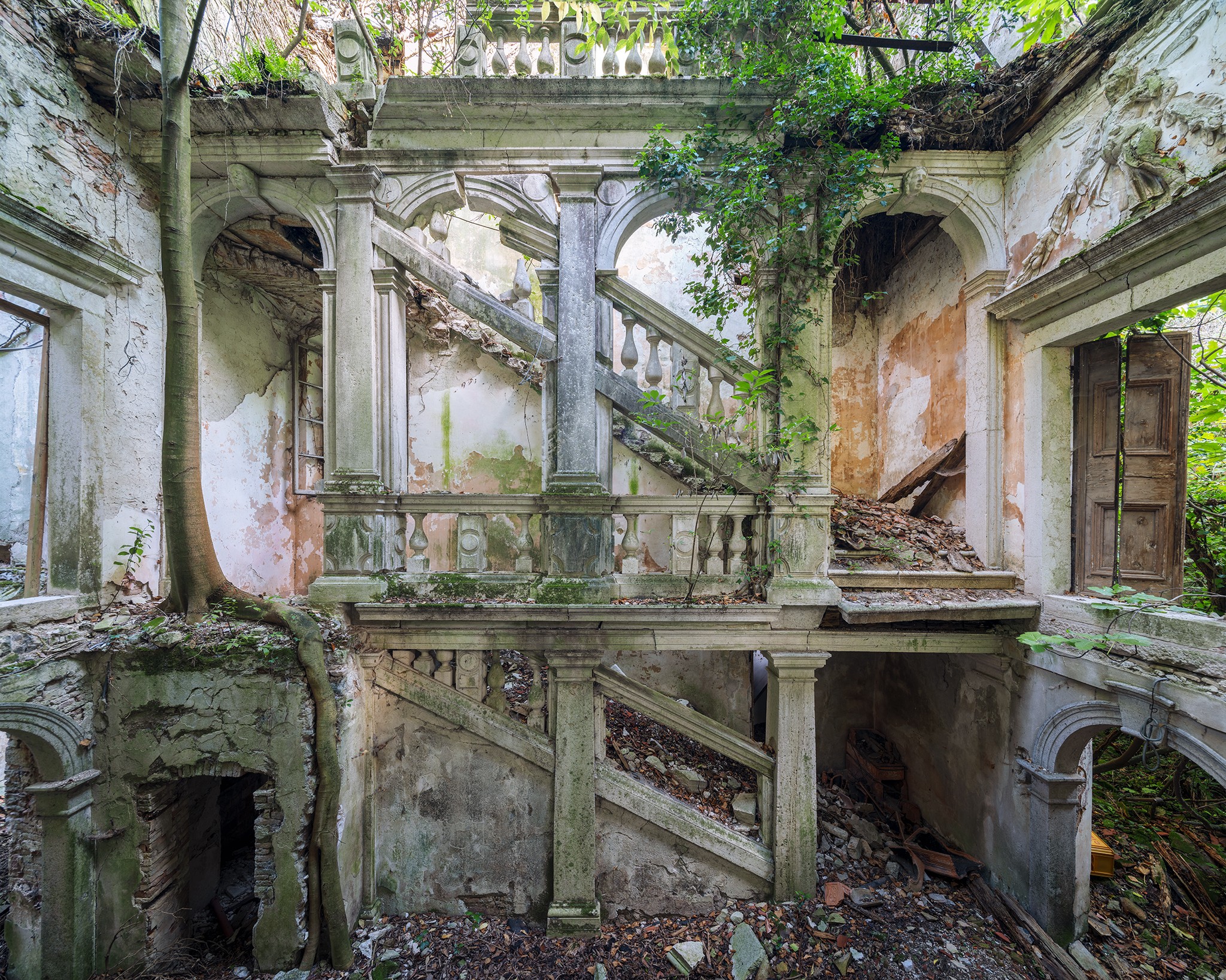 Ruined, overgrown staircase in an abandoned building with crumbling walls and vegetation.