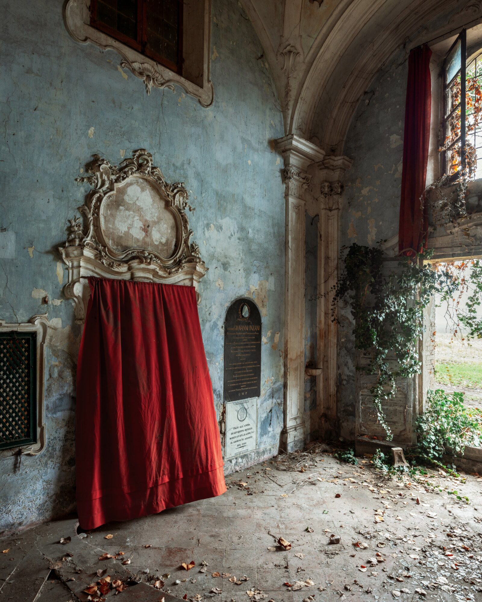 Interior of an old building with a red curtain covering a decorative frame, a window with sunlight, and plants near the window.