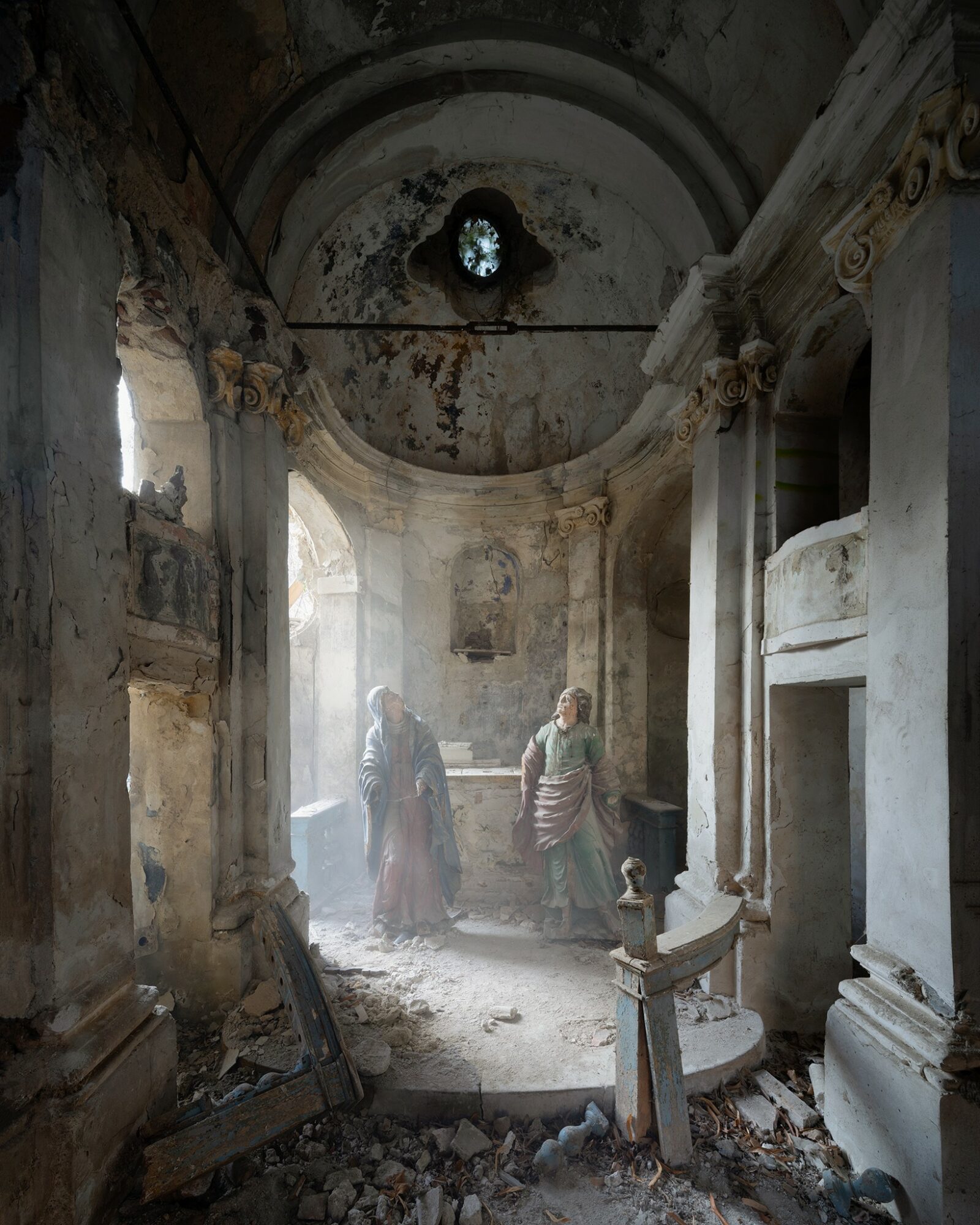 Interior of a damaged, abandoned building with two people standing inside, sunlight streaming through broken windows, debris on the floor.