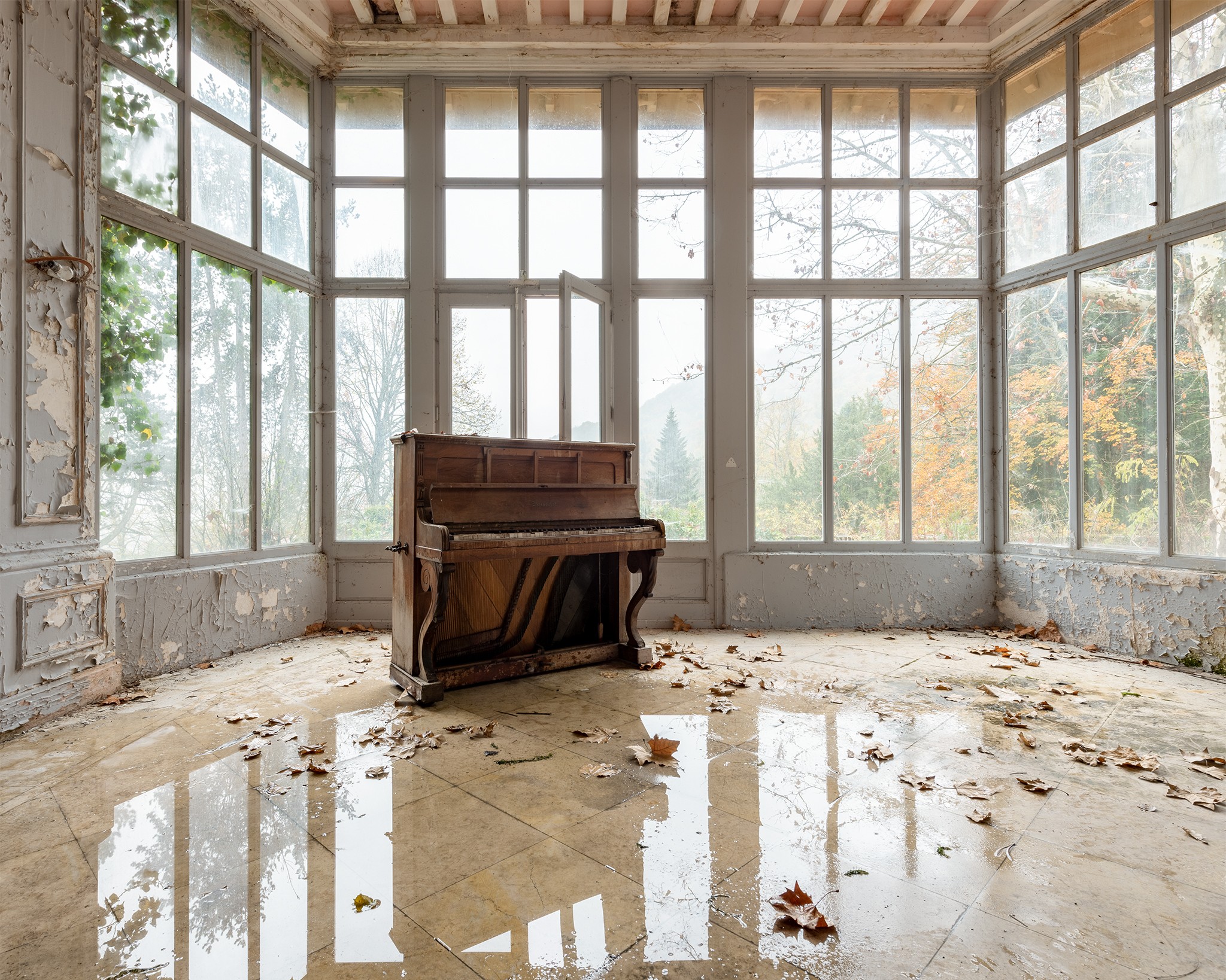 Old piano in a sunlit, abandoned glass room with debris on the floor.