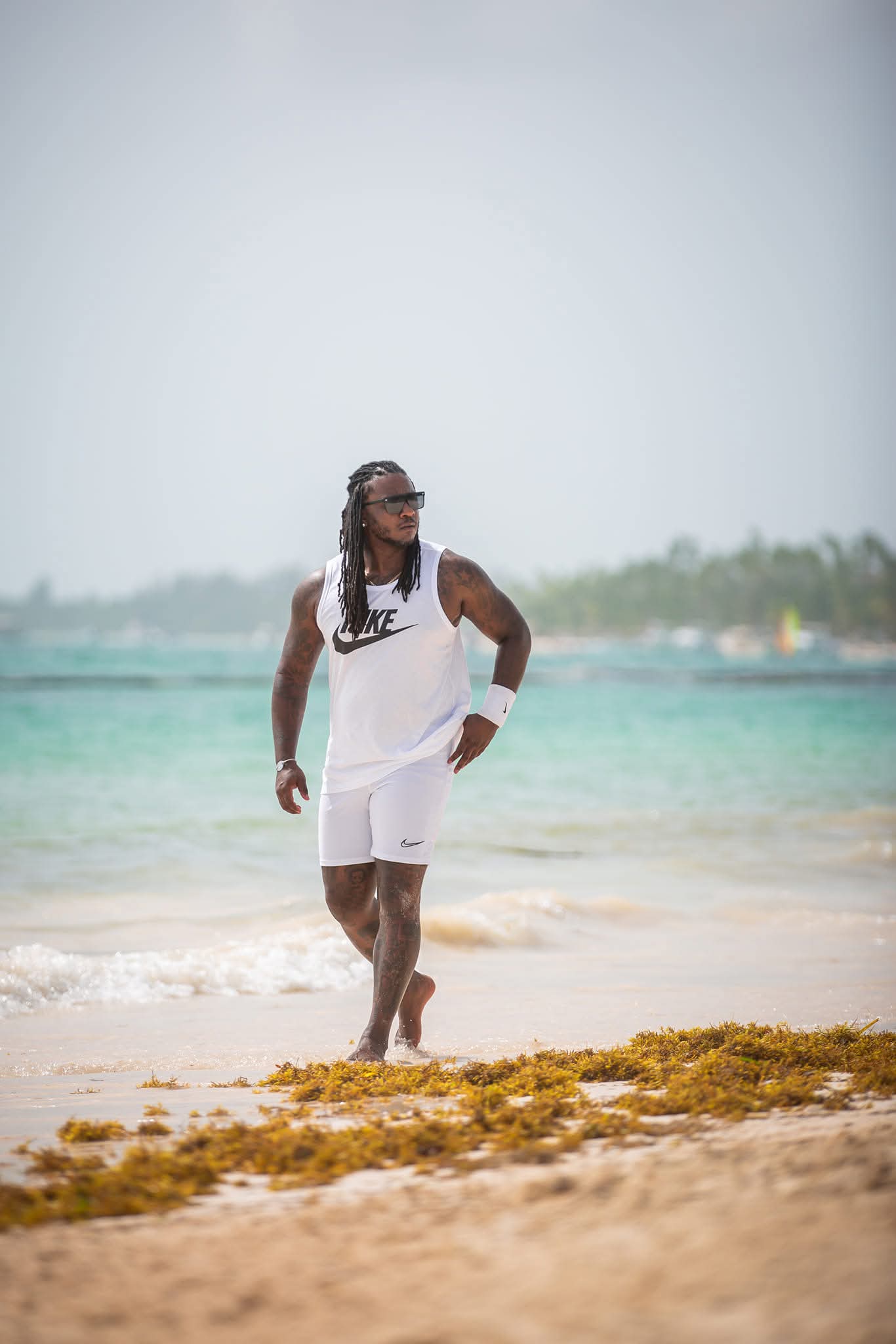 Man with long hair and sunglasses walking on beach in white sleeveless shirt and shorts, ocean and sky in background.