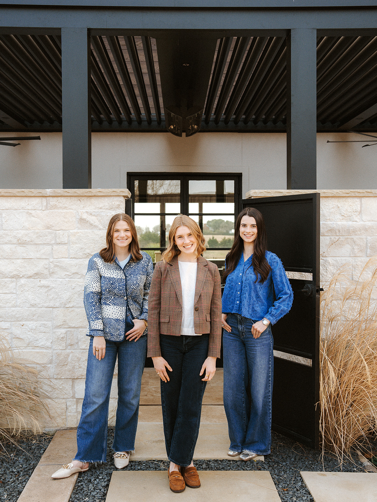 Three women standing outdoors in front of a stone wall and black gate, smiling, with a modern building in the background.