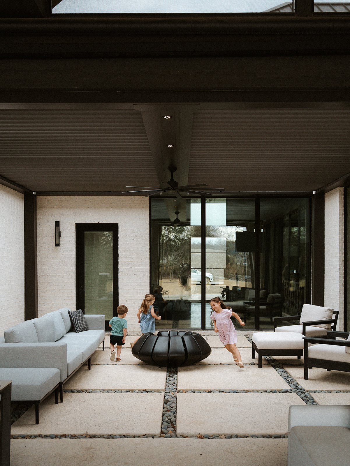 Four children playing on a covered outdoor patio with modern furniture and large glass doors, daytime.