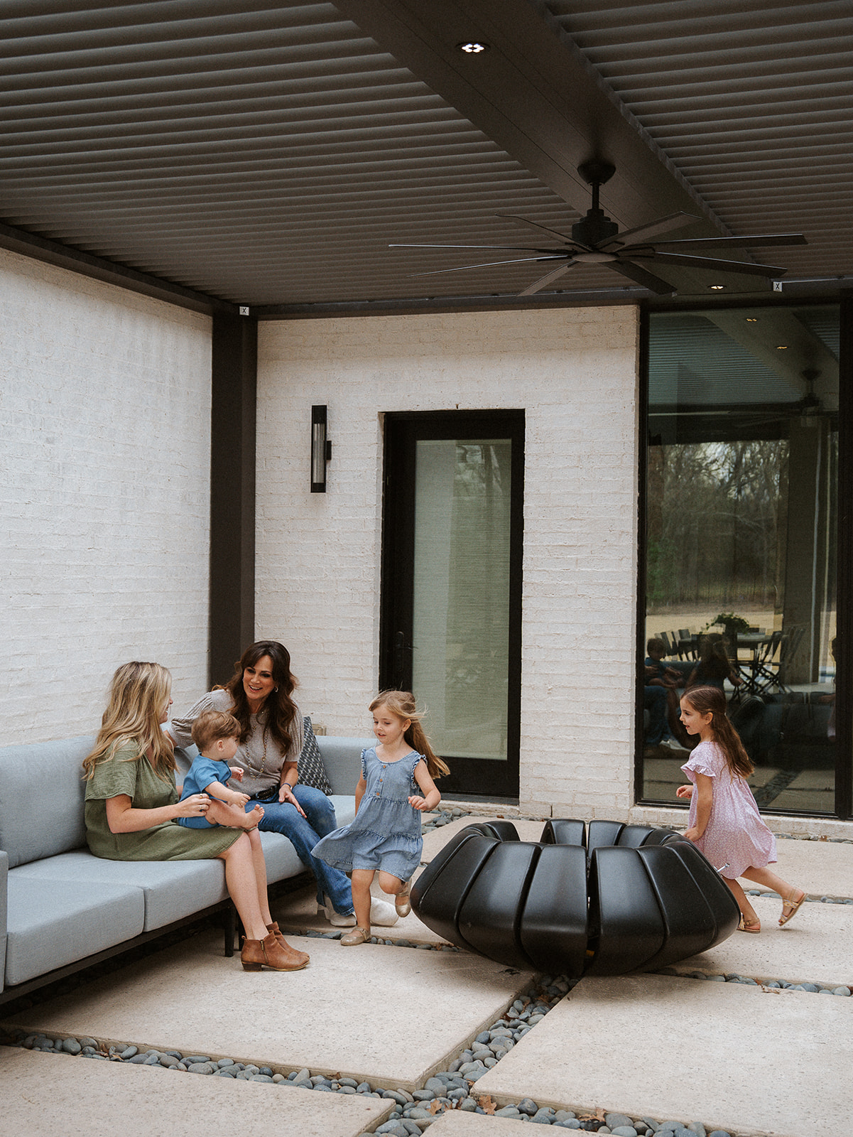 Four children and an adult sitting and playing on a sofa in a modern outdoor patio area with a large black fire pit.