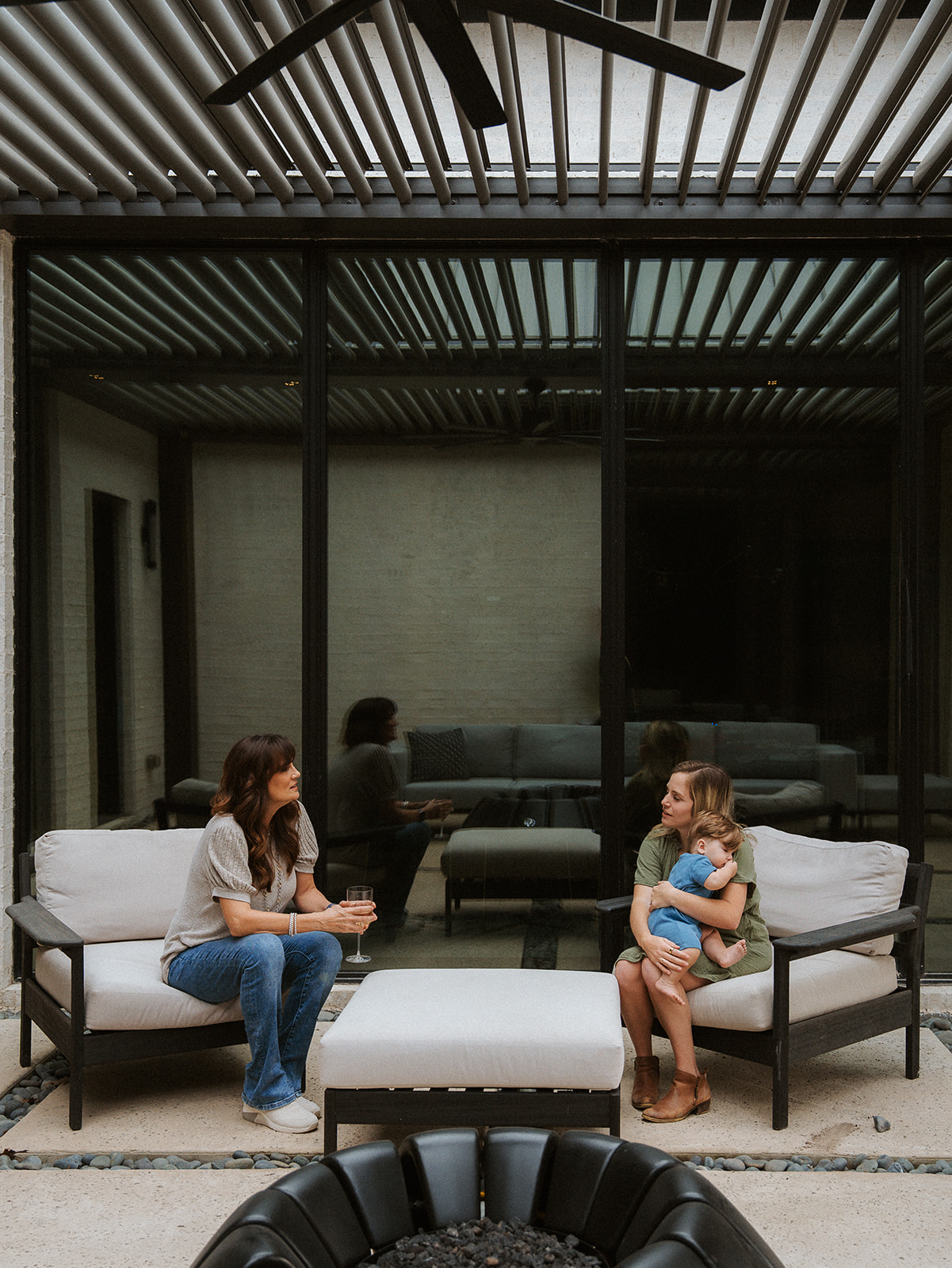 Two women sitting on outdoor sofas with a small child, in a modern patio area with a glass wall and wooden ceiling.