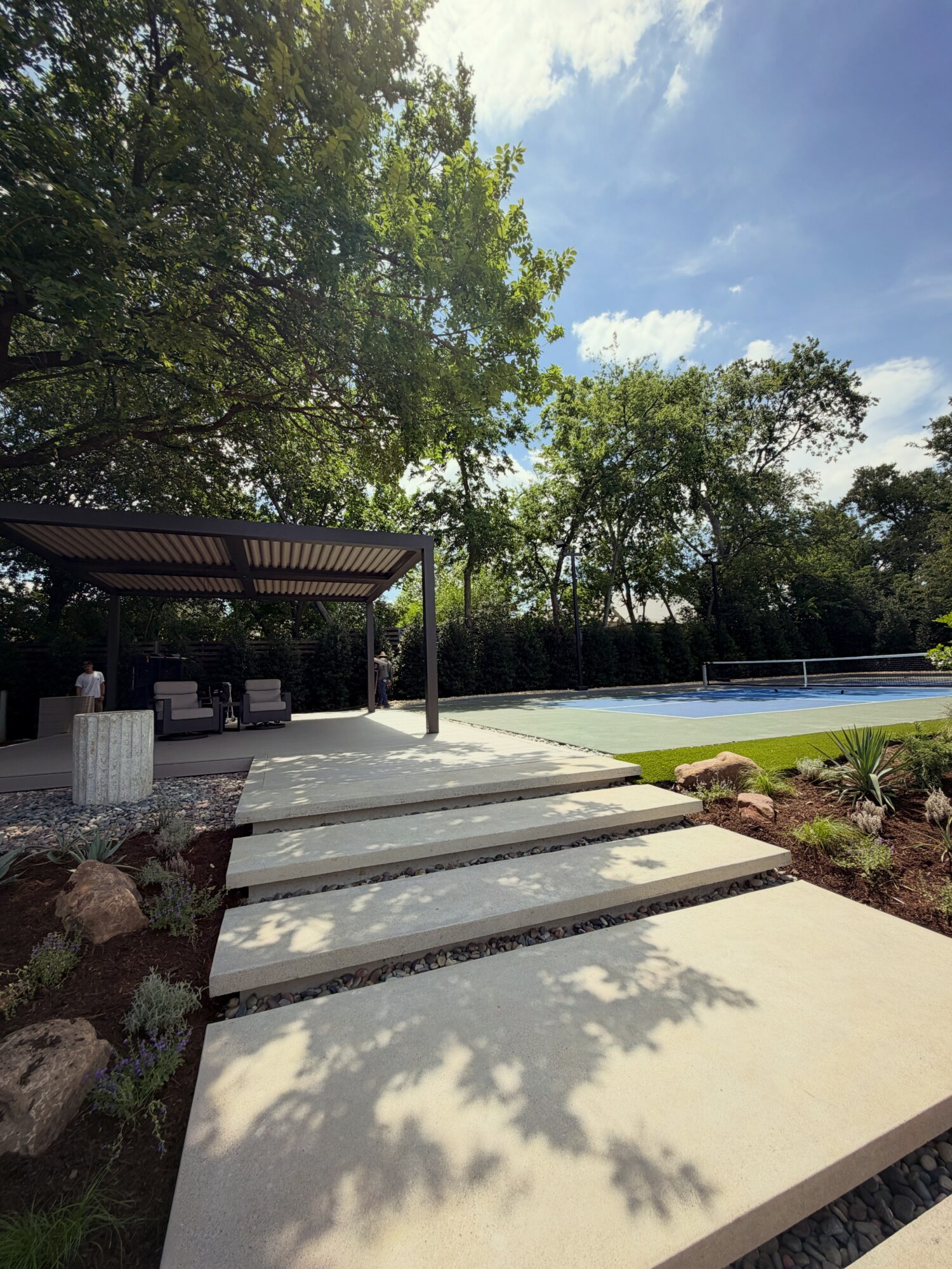 Concrete pathway leading to a pool area with trees and a shaded seating area in the background.