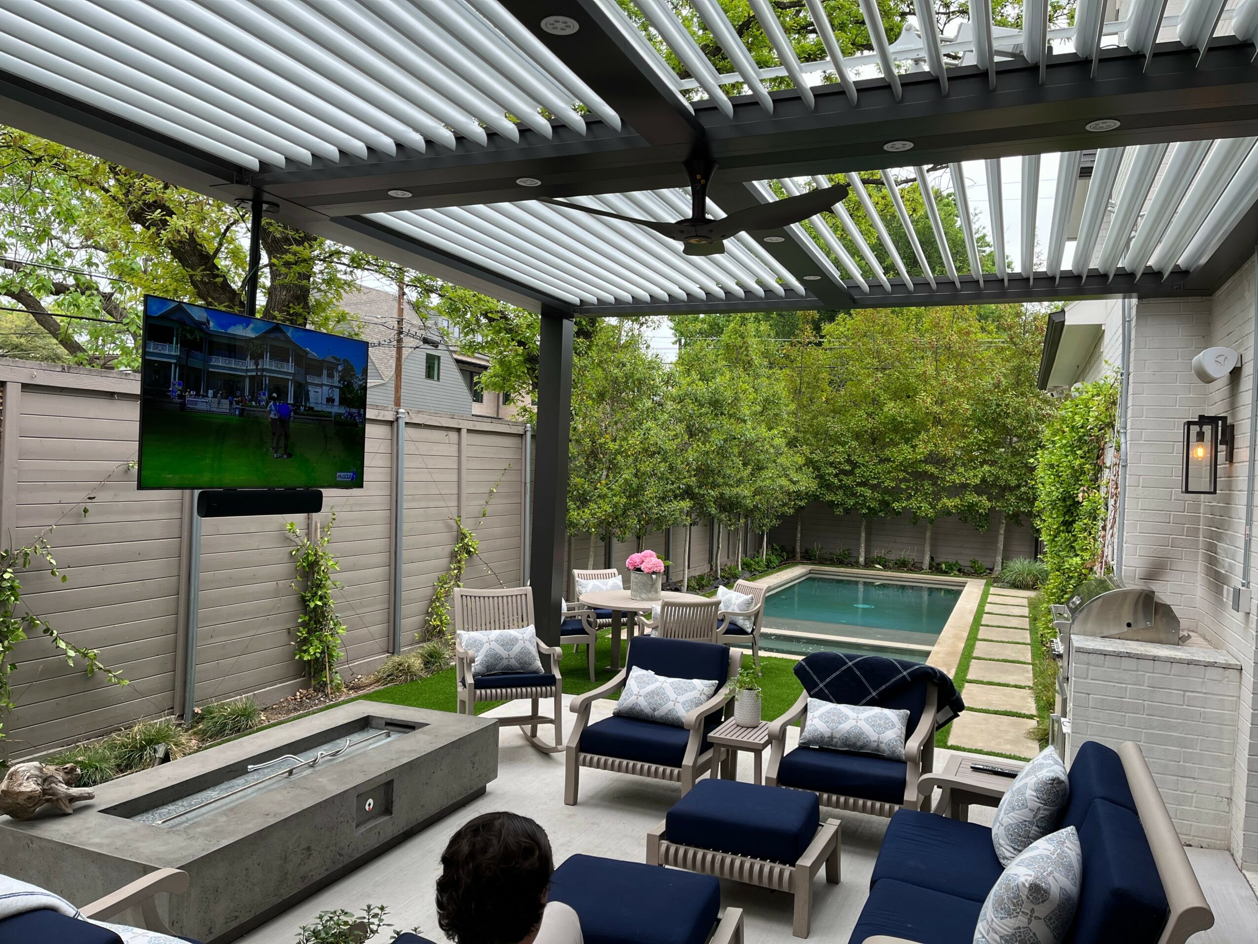 Outdoor patio with seating area, pool, and greenery, under a pergola with a TV mounted on the wall.