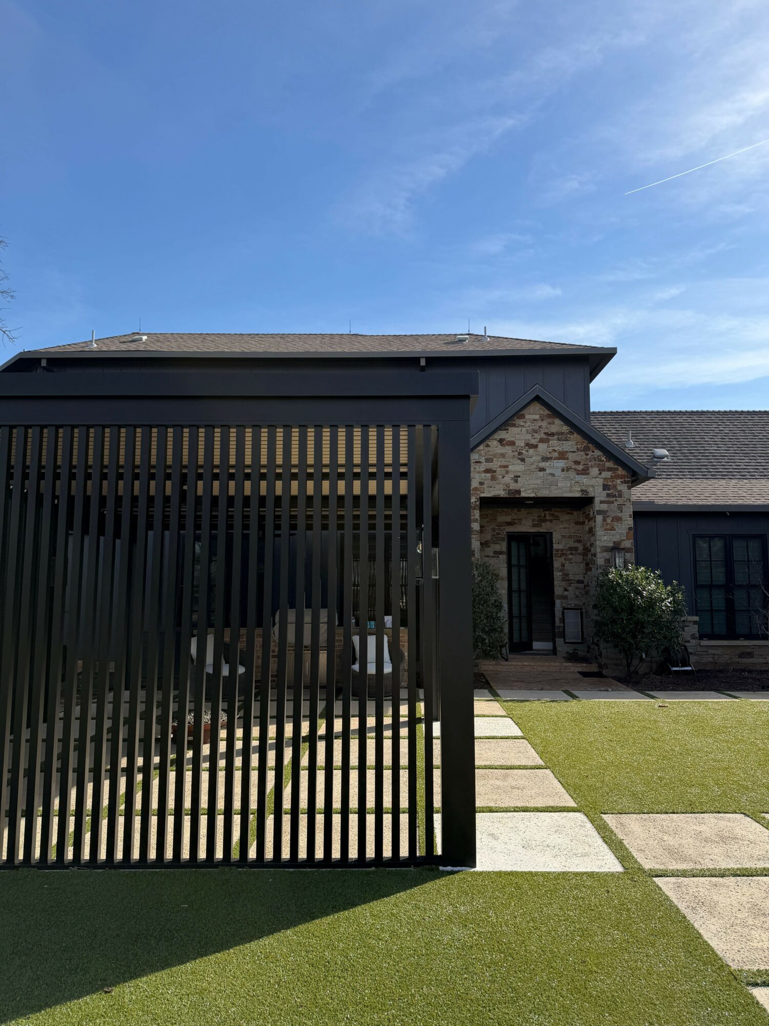 A modern house with a black slatted gate, stone walls, and a green lawn under a blue sky.