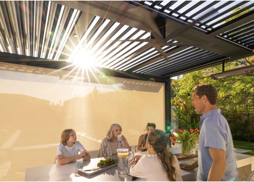 Group of five people sitting at a table outdoors, sunlight shining through a slatted roof, greenery in background.