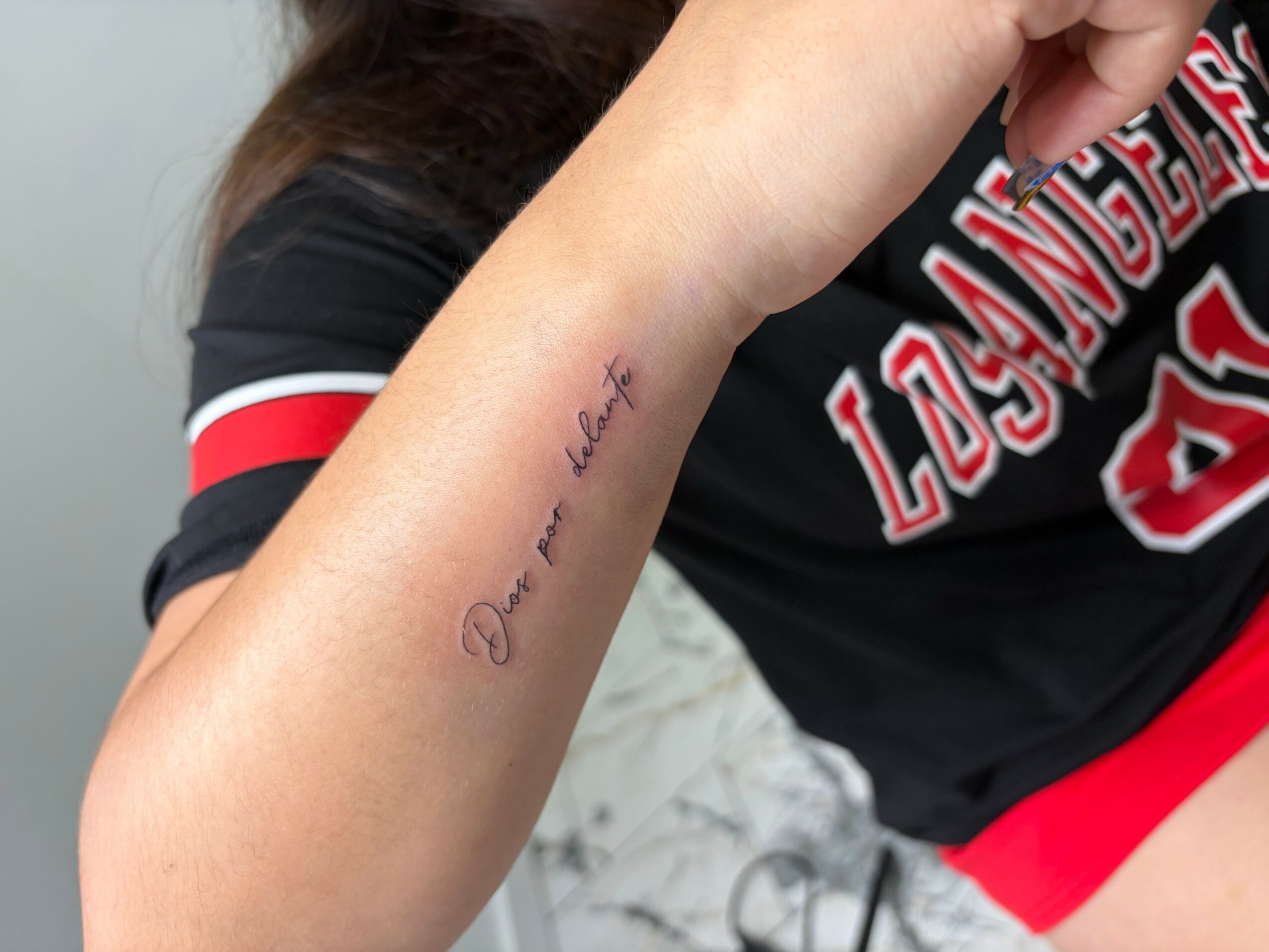 Person with a tattoo on their forearm, wearing a black shirt with red and white lettering, holding a pen near their face.