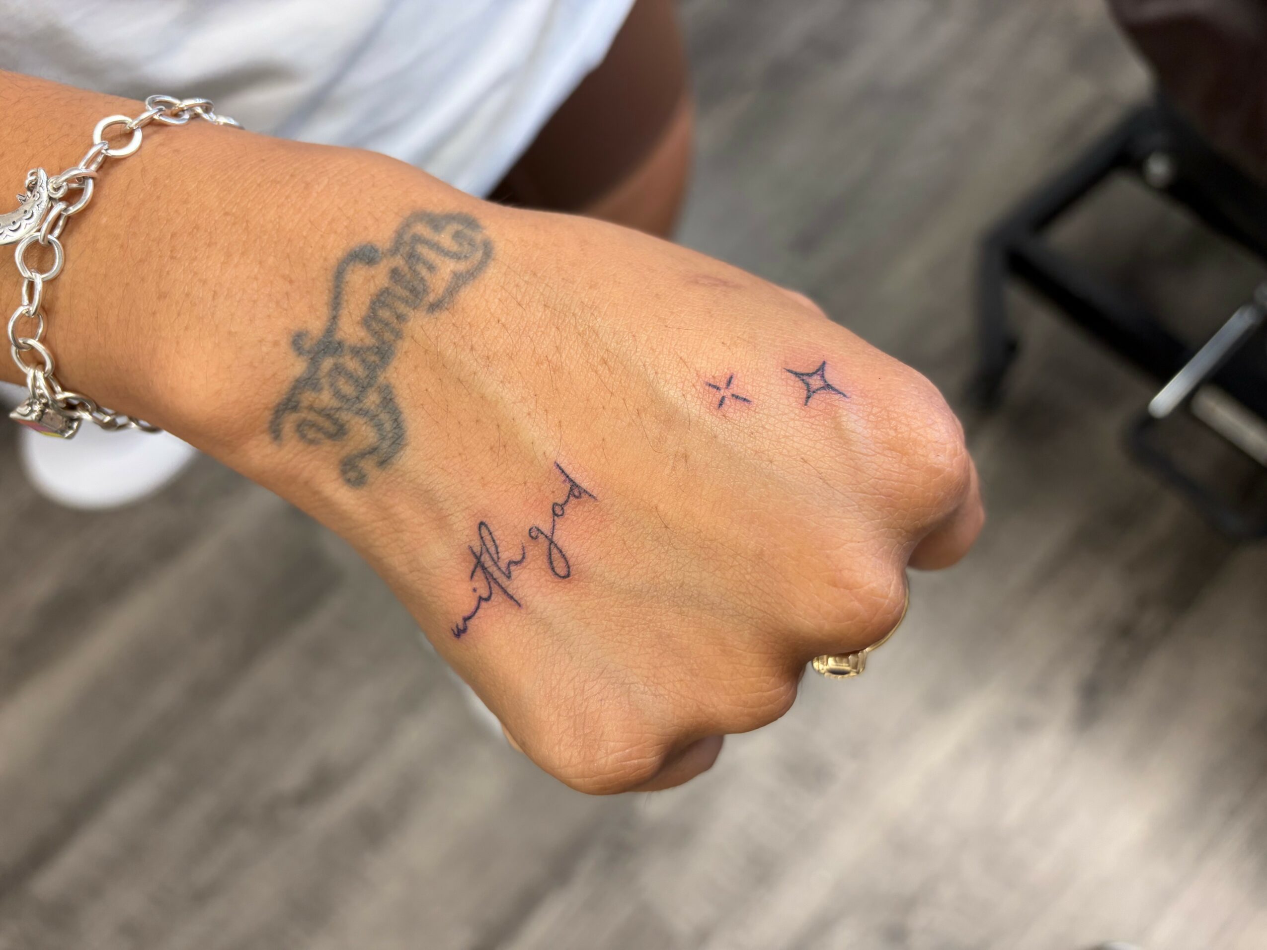 Fist with tattoos and a bracelet, background shows wooden floor and part of a chair, no text visible.