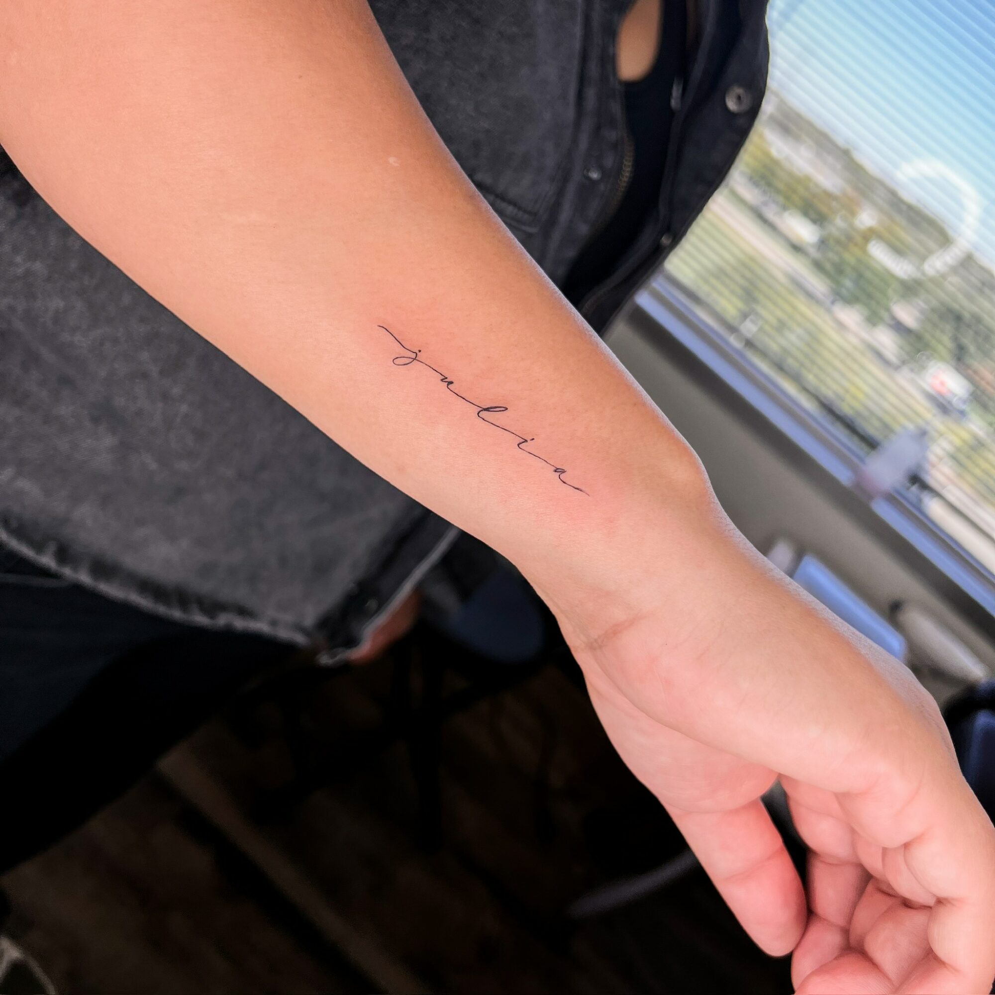Person's forearm with a small, delicate tattoo of a handwritten word, in a well-lit room with a window view.