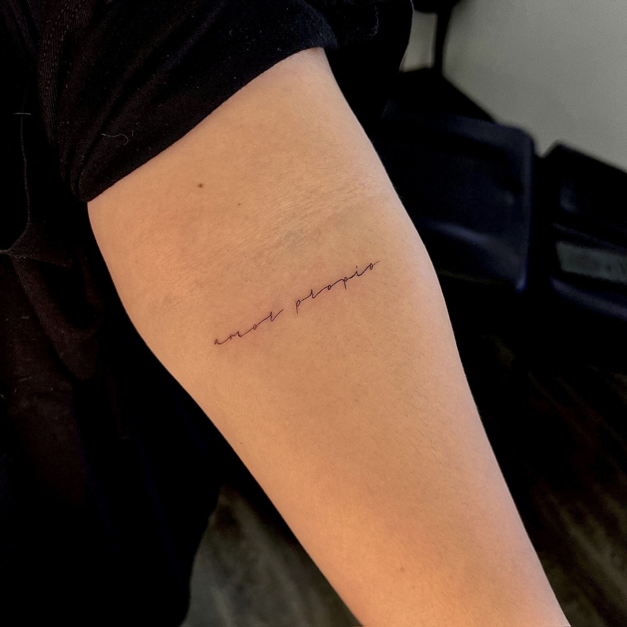Person's upper arm with a small, handwritten tattoo in black ink, partially visible, against a dark background.