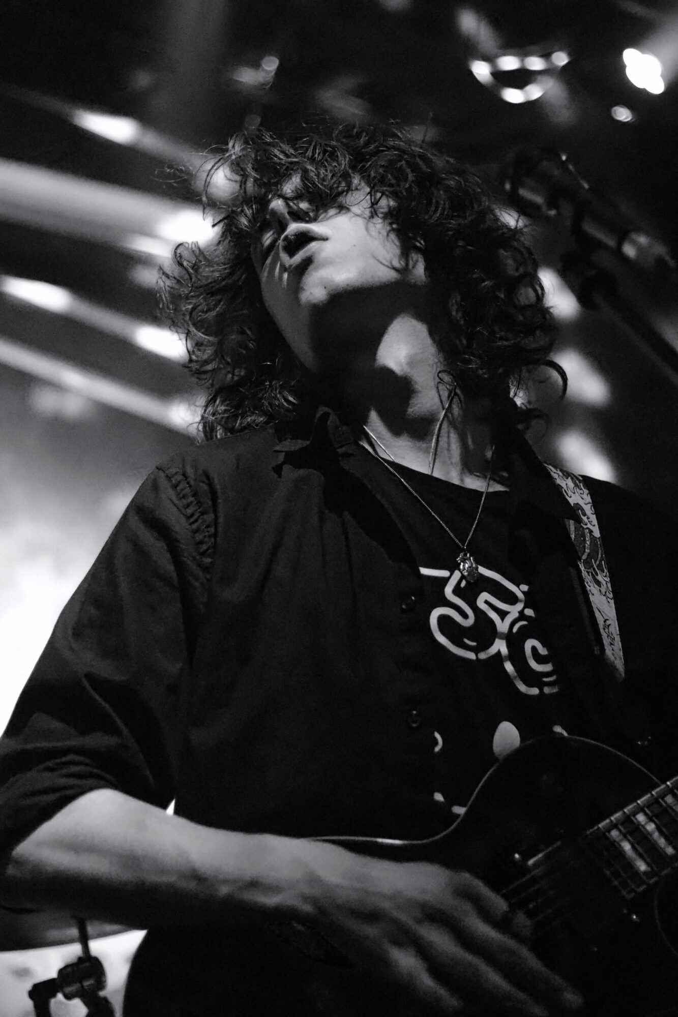 Person with curly hair playing guitar on stage, head tilted back, in black and white.