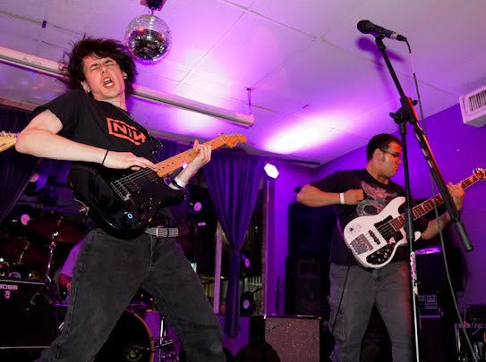 Two musicians perform on stage with guitars, purple lighting, and a disco ball overhead.