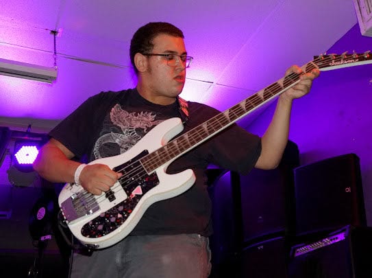 Person playing an electric guitar on stage with purple lighting, wearing glasses and a dark T-shirt.