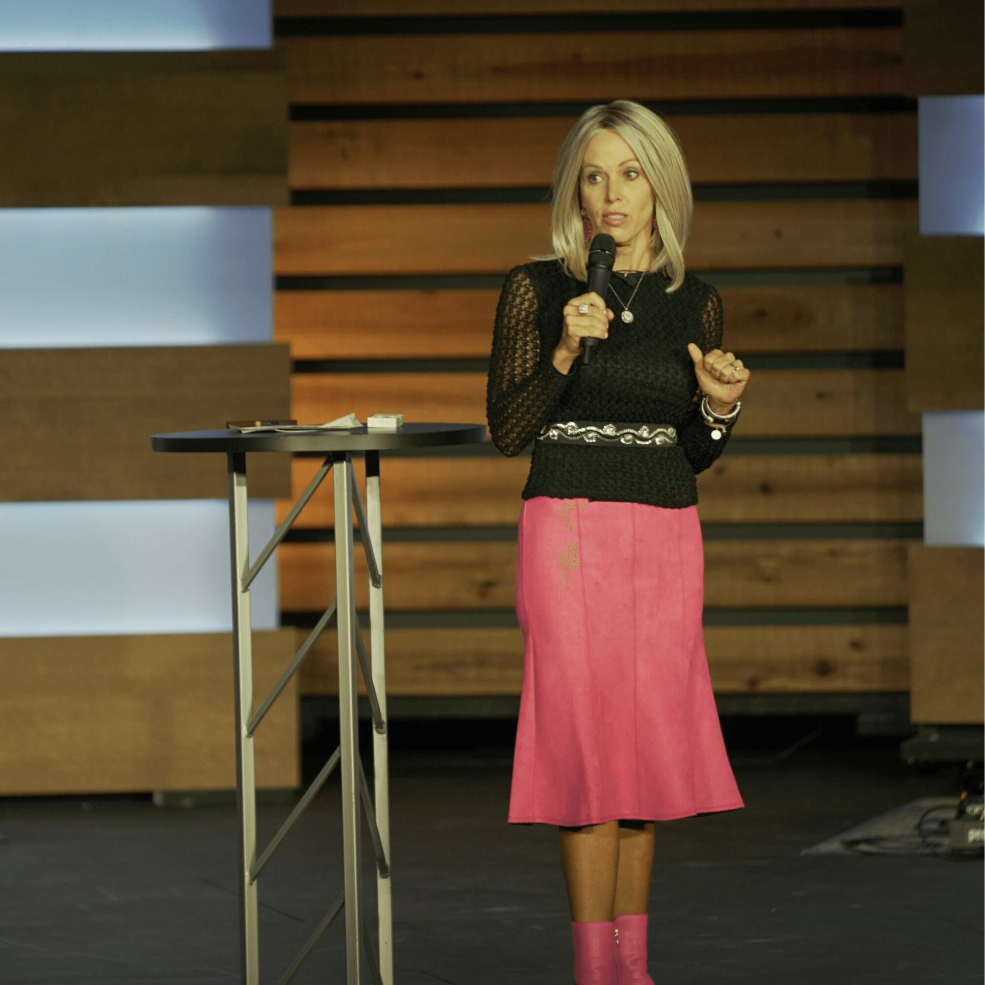 Woman with blonde hair speaking into a microphone on stage, wearing a black top and pink skirt, with a wooden background.