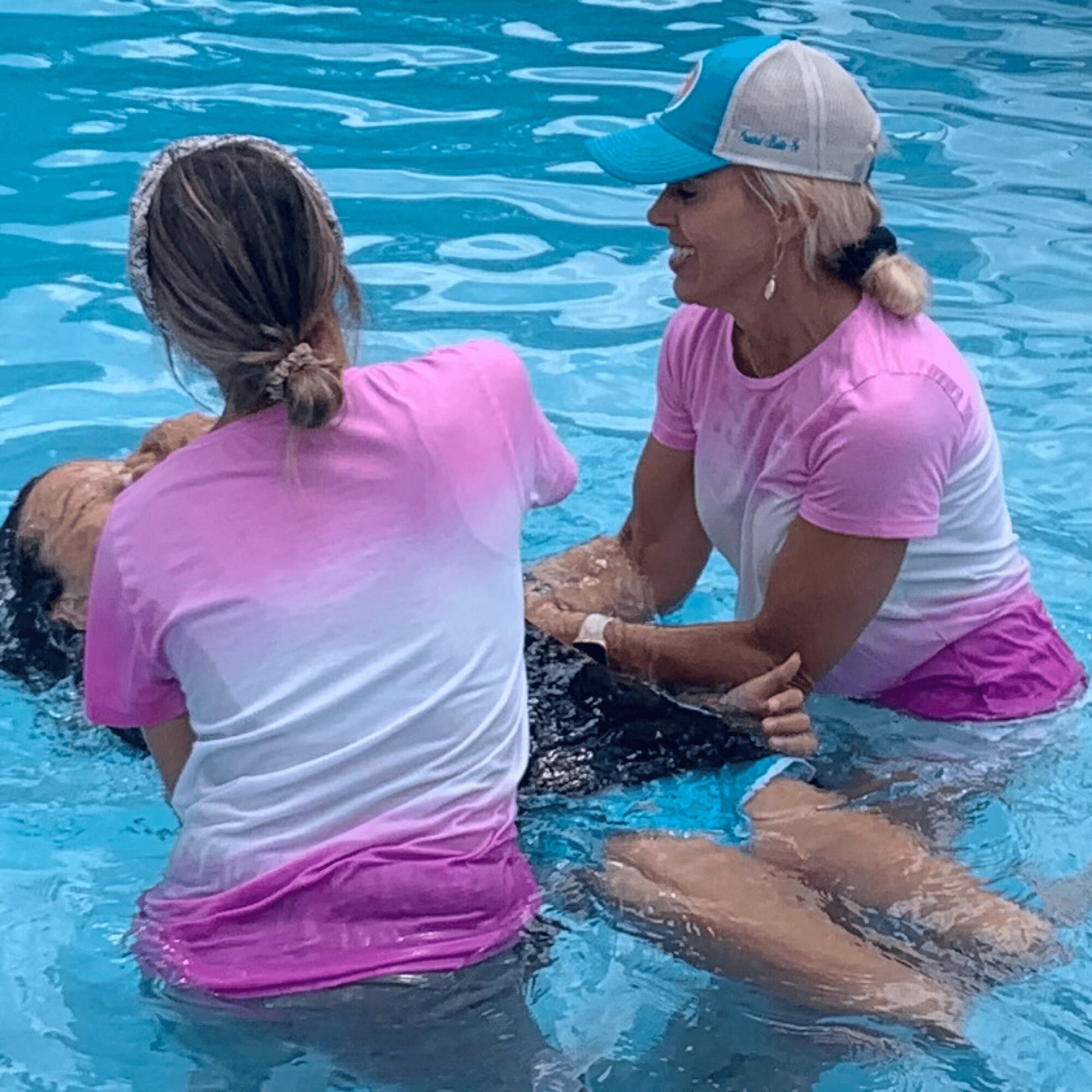 Two women in pink and white shirts assist a person in a swimming pool, holding their arms. One woman wears a cap.