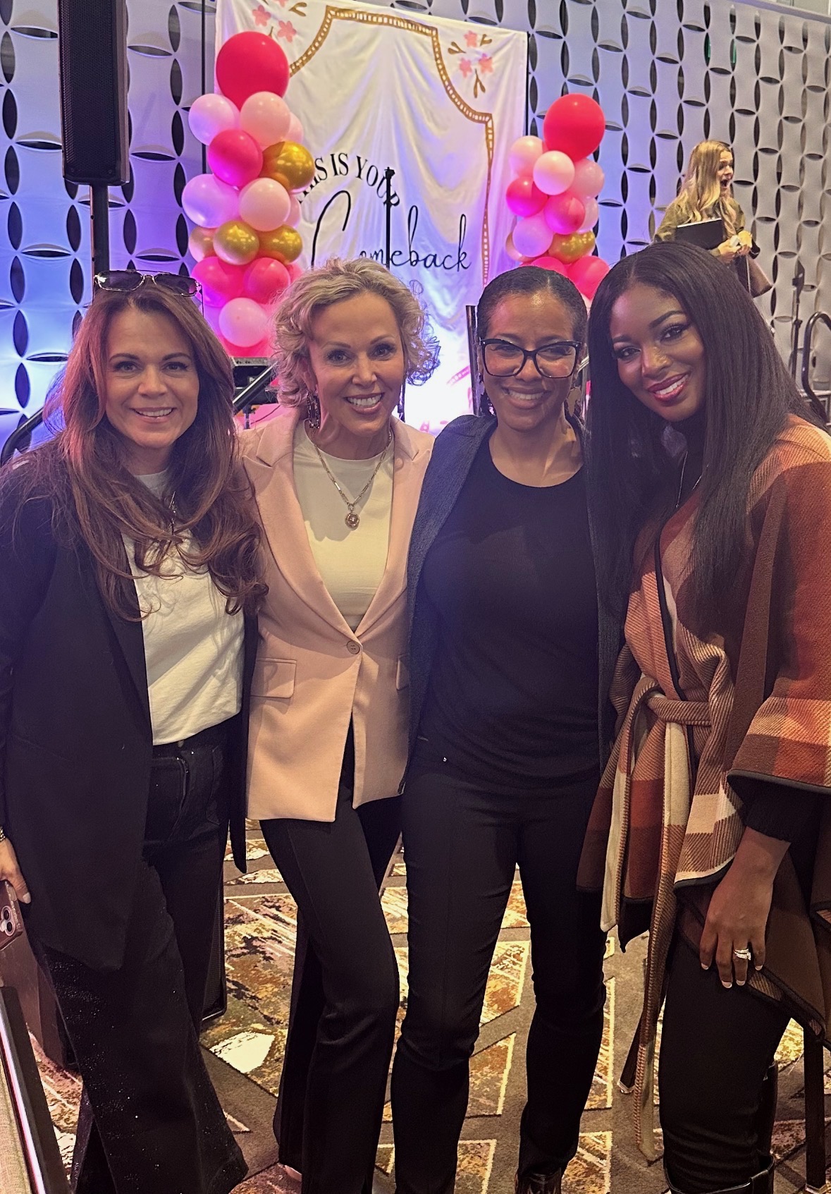 Four women standing together at an event with pink and gold balloons and a decorated backdrop.