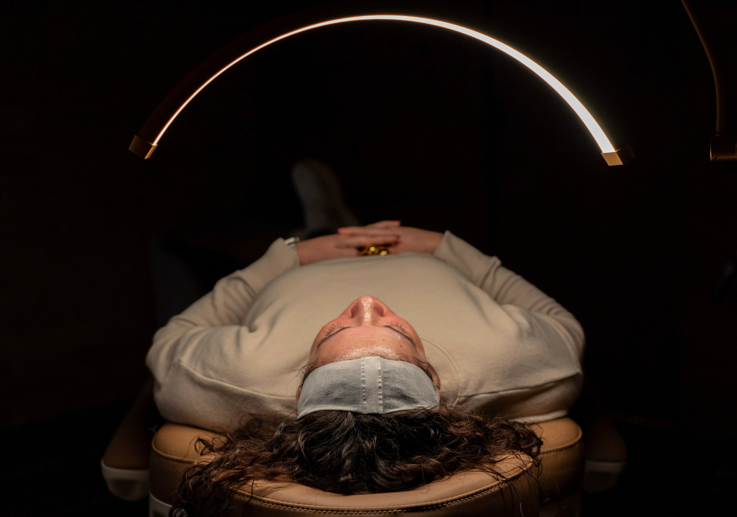 Person lying on a table with head tilted back, illuminated by a curved light above, in a dark room.