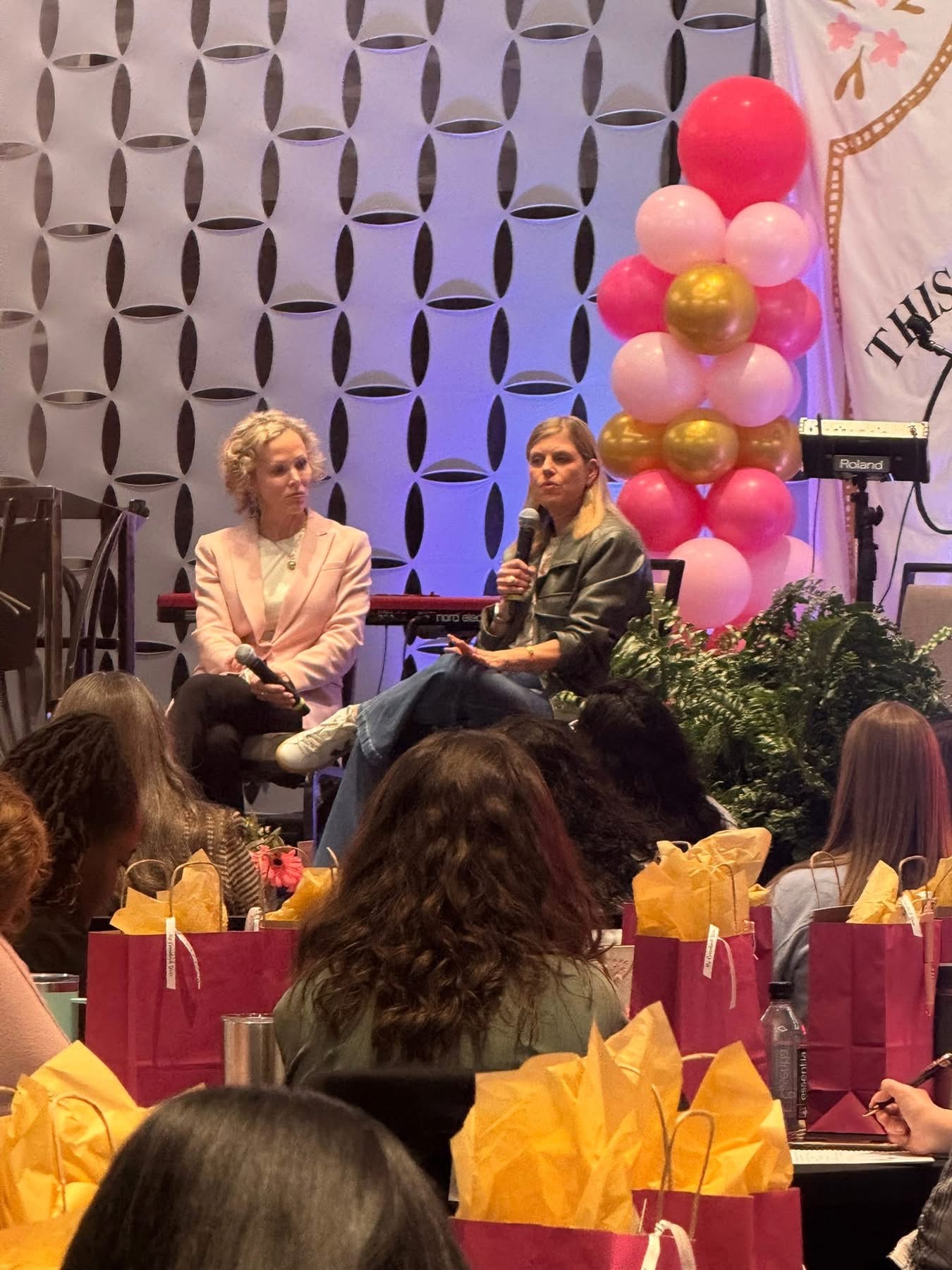 Two women sit on stage in front of an audience, with a pink and gold balloon decoration behind them, and a patterned backdrop.