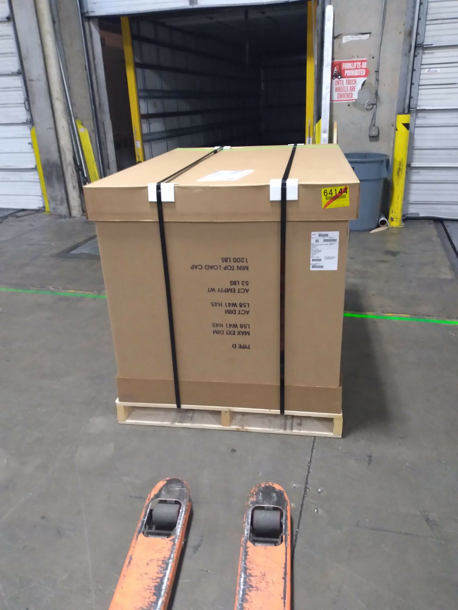 Large cardboard box on a pallet inside a warehouse, with a forklift nearby, and warehouse equipment in the background.
