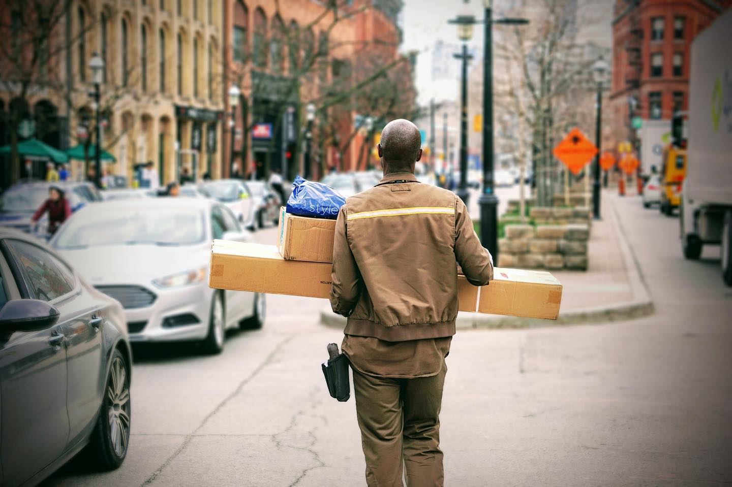 Person walking on city sidewalk carrying a long box, with cars and buildings in the background.