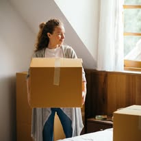 Young woman holding a cardboard box in a sunlit room with a window and curtains.