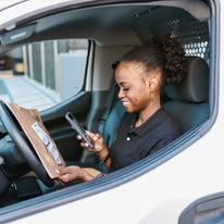 Woman sitting in car, smiling, looking at her phone, holding a package, with a window showing an urban background.