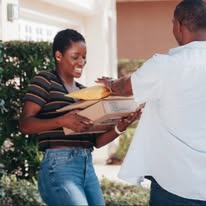 Person receiving a package from another person outdoors, smiling, with greenery and a building in the background.