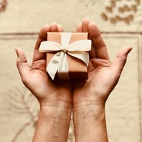 Person holding a small gift box with a white ribbon bow, on a wooden surface.